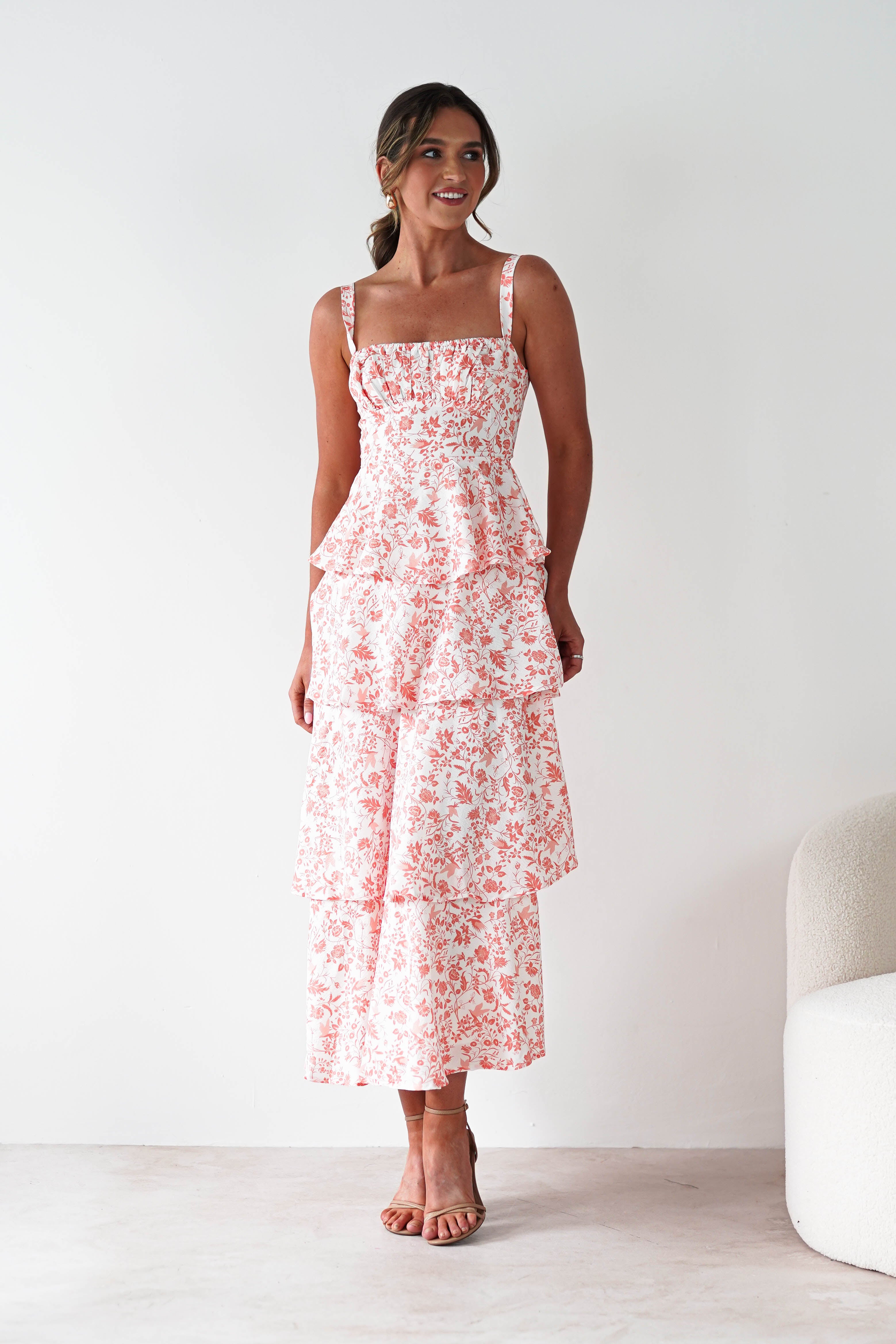 A woman stands indoors wearing a sleeveless, pink and white floral tiered maxi dress with ruffle detail. Perfect for wedding guests, she smiles with her hair tied back, posed next to a white curved chair against a plain white wall.