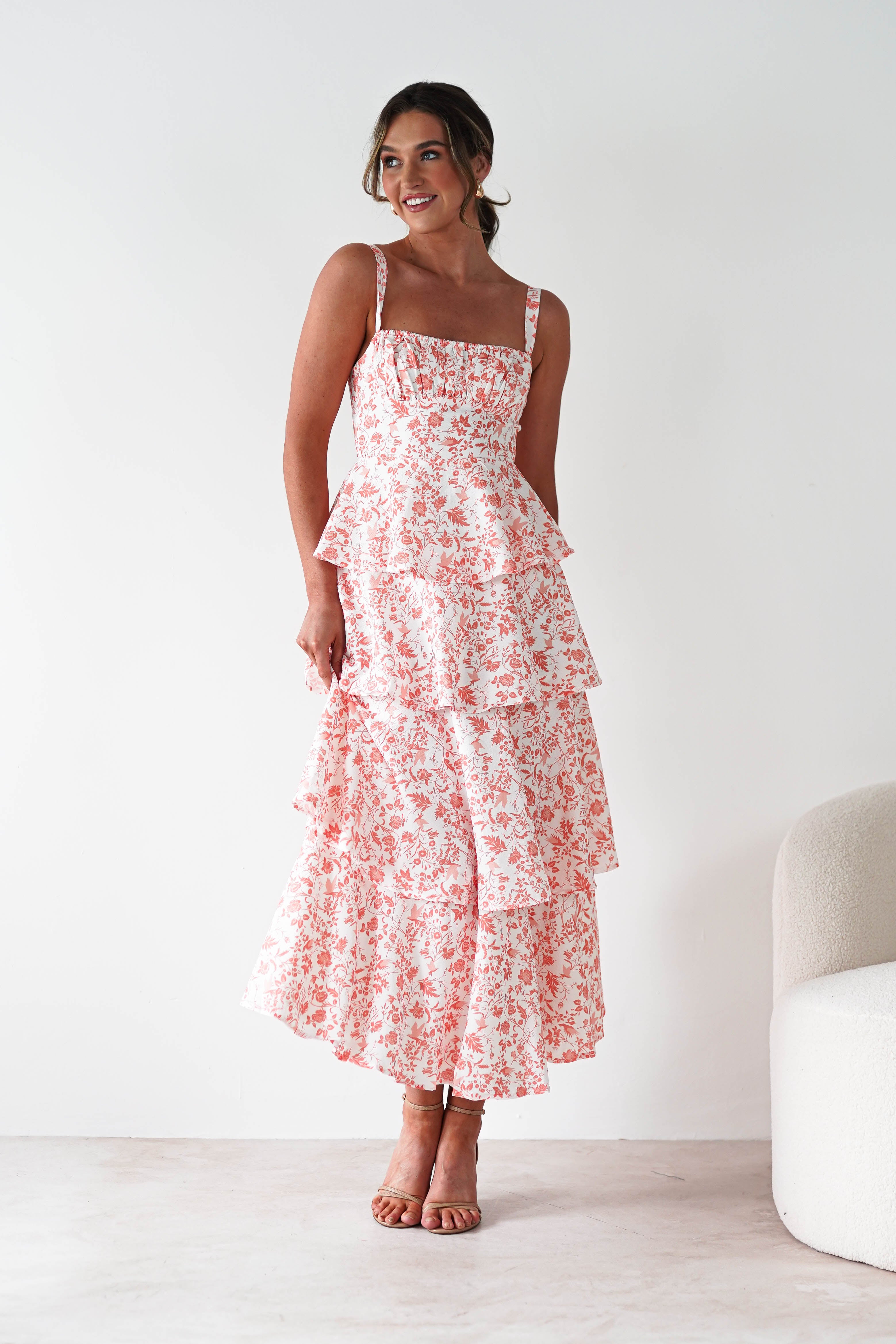 A woman stands indoors, smiling in a summer orange print maxi dress with ruffle detail. Her hair is tied back, and she poses next to a light-colored, curved chair—perfect attire for wedding guests.