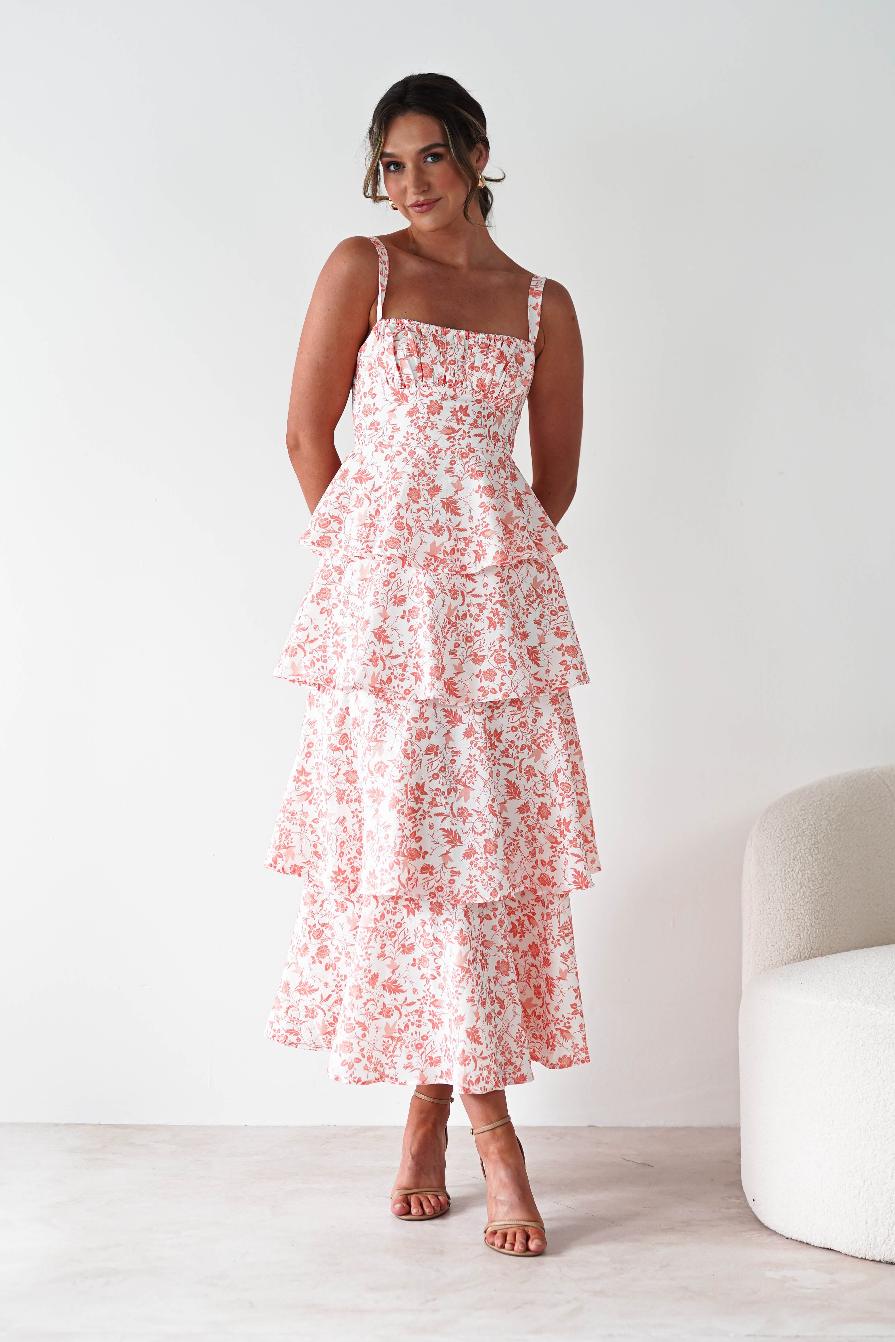 Woman standing in a sleeveless, tiered, ankle-length dress with a pink floral pattern and subtle ruffle detail, looking slightly to the side. Her hands are behind her back. The minimal setting is perfect for wedding guests or summer gatherings.