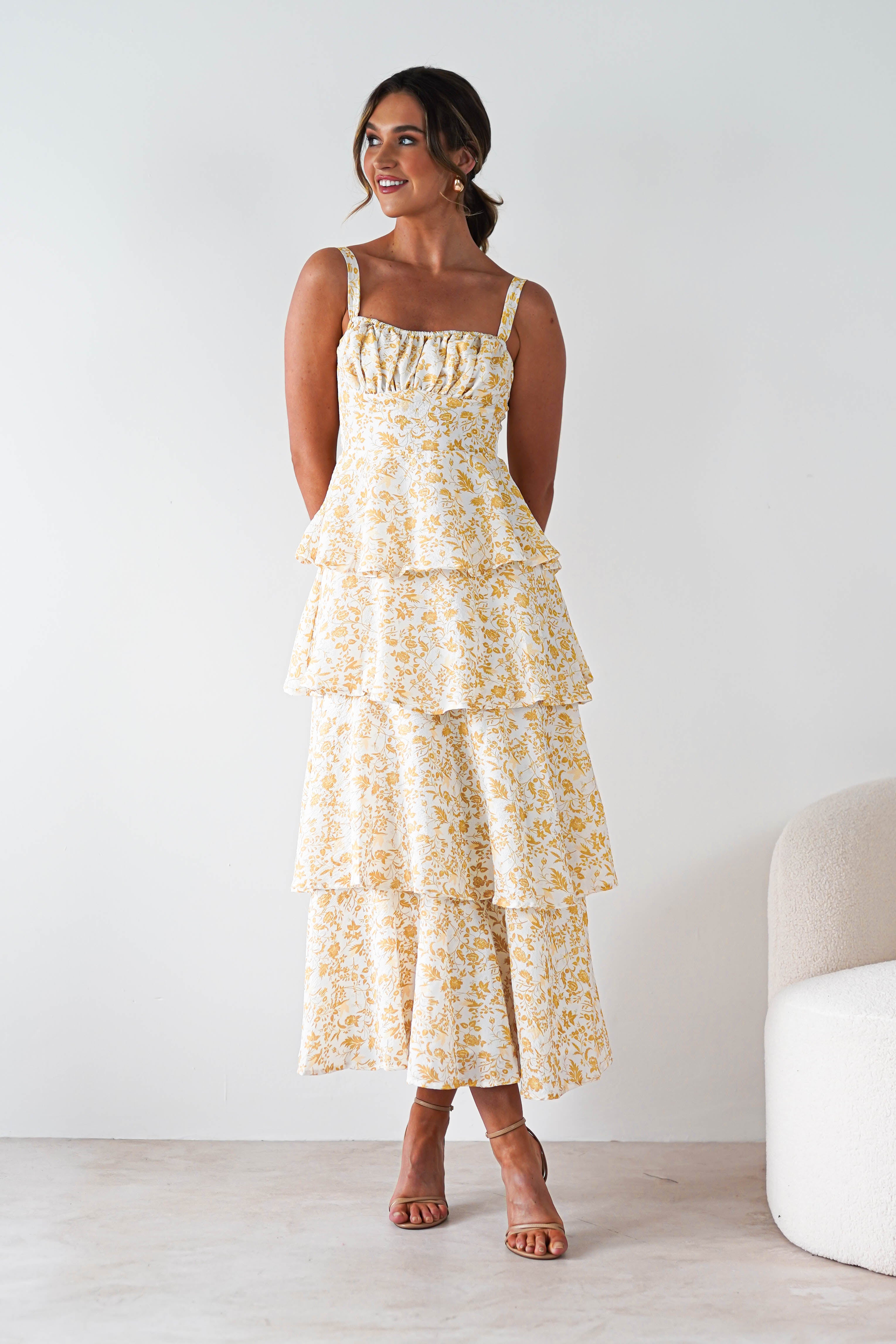 A woman wearing a yellow print maxi dress with ruffle detail stands indoors, smiling and looking to the side. Her hair is styled up, and she pairs the look with tan heeled sandals—perfect for summer events.