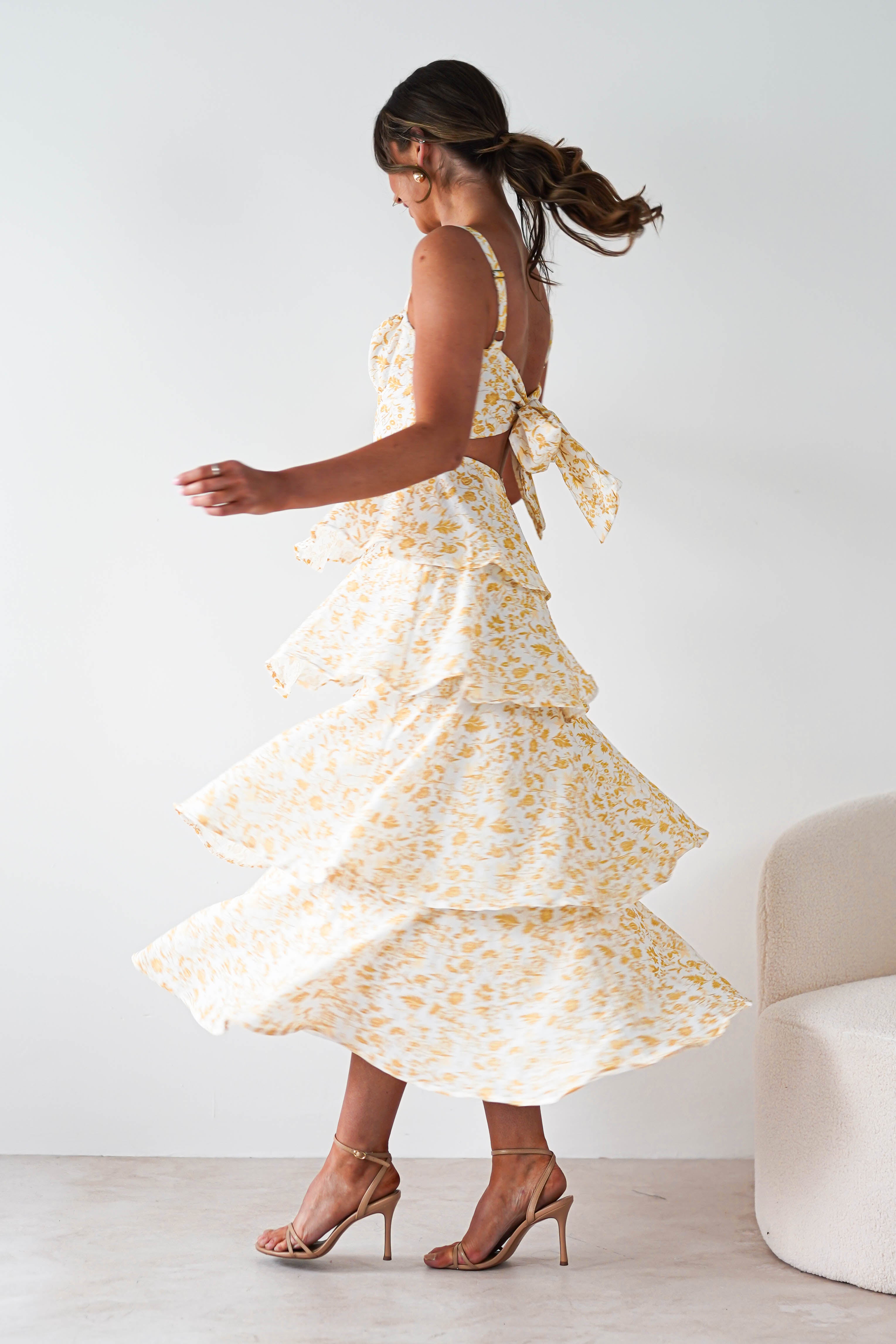 A woman twirls in a sleeveless yellow print maxi dress with ruffle detail, open back, and bow—perfect for summer events. She wears high-heeled sandals, her long brown hair in a ponytail, beside a light-colored sofa against a plain background.