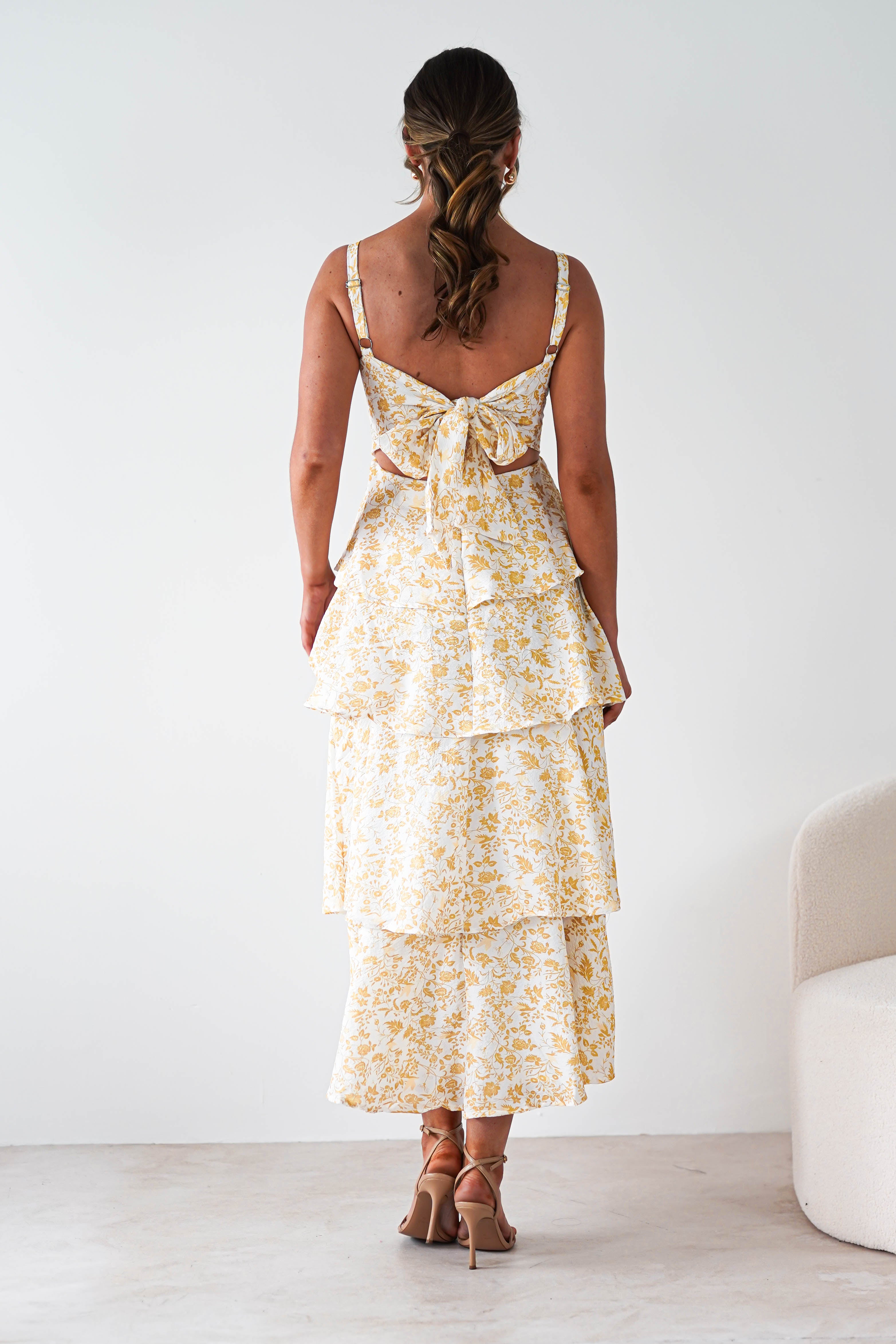 A woman stands facing away, wearing a sleeveless, yellow print maxi dress with ruffle detail and a large bow at the back. Her hair is styled in loose waves, and she is wearing heeled sandals. The background is minimal and light.