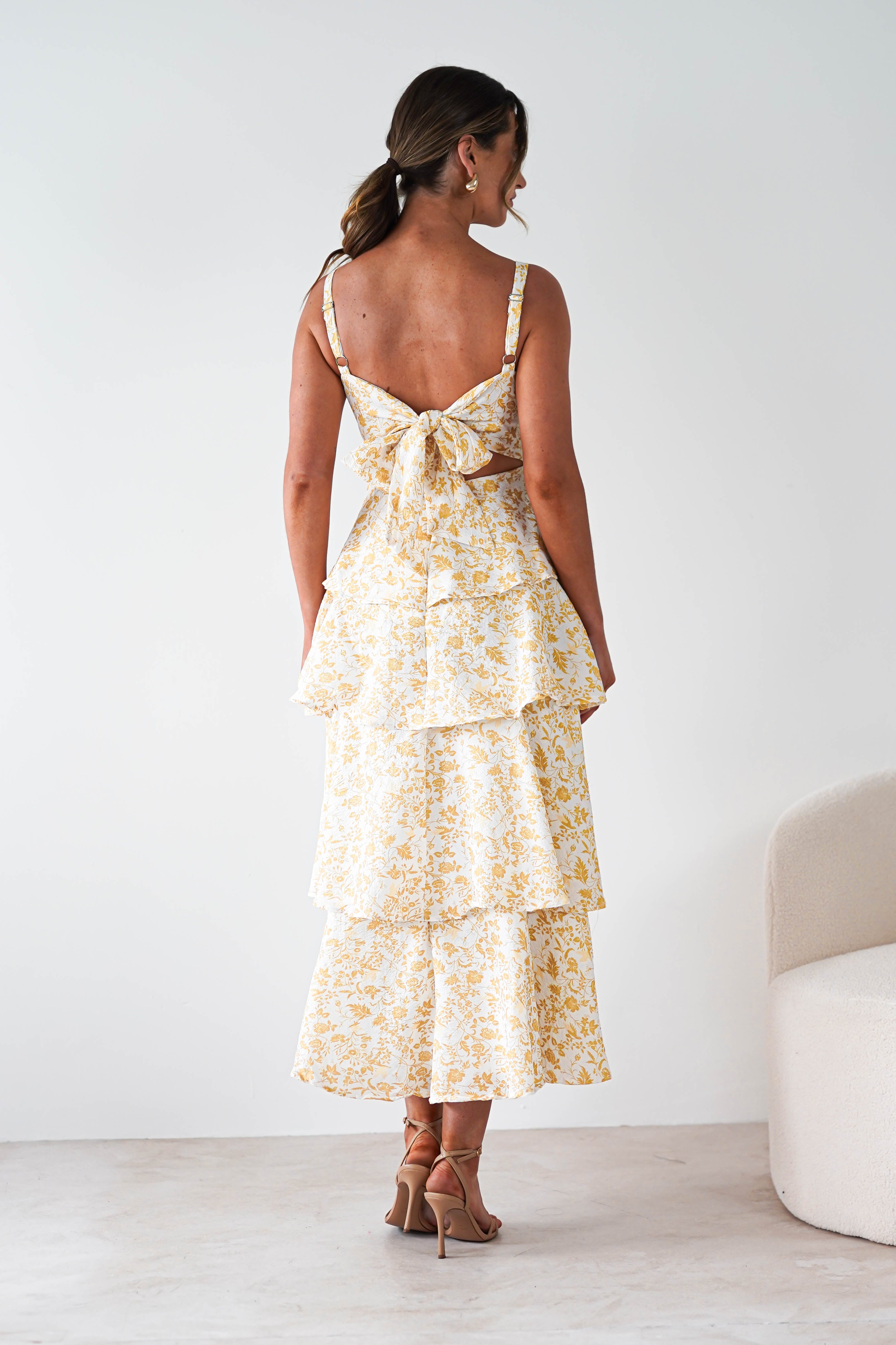 A woman stands facing away in a yellow print maxi dress with ruffle detail, featuring tiers and a large bow at the back. With wavy hair and heels, she’s ready for summer events near a light-colored chair.