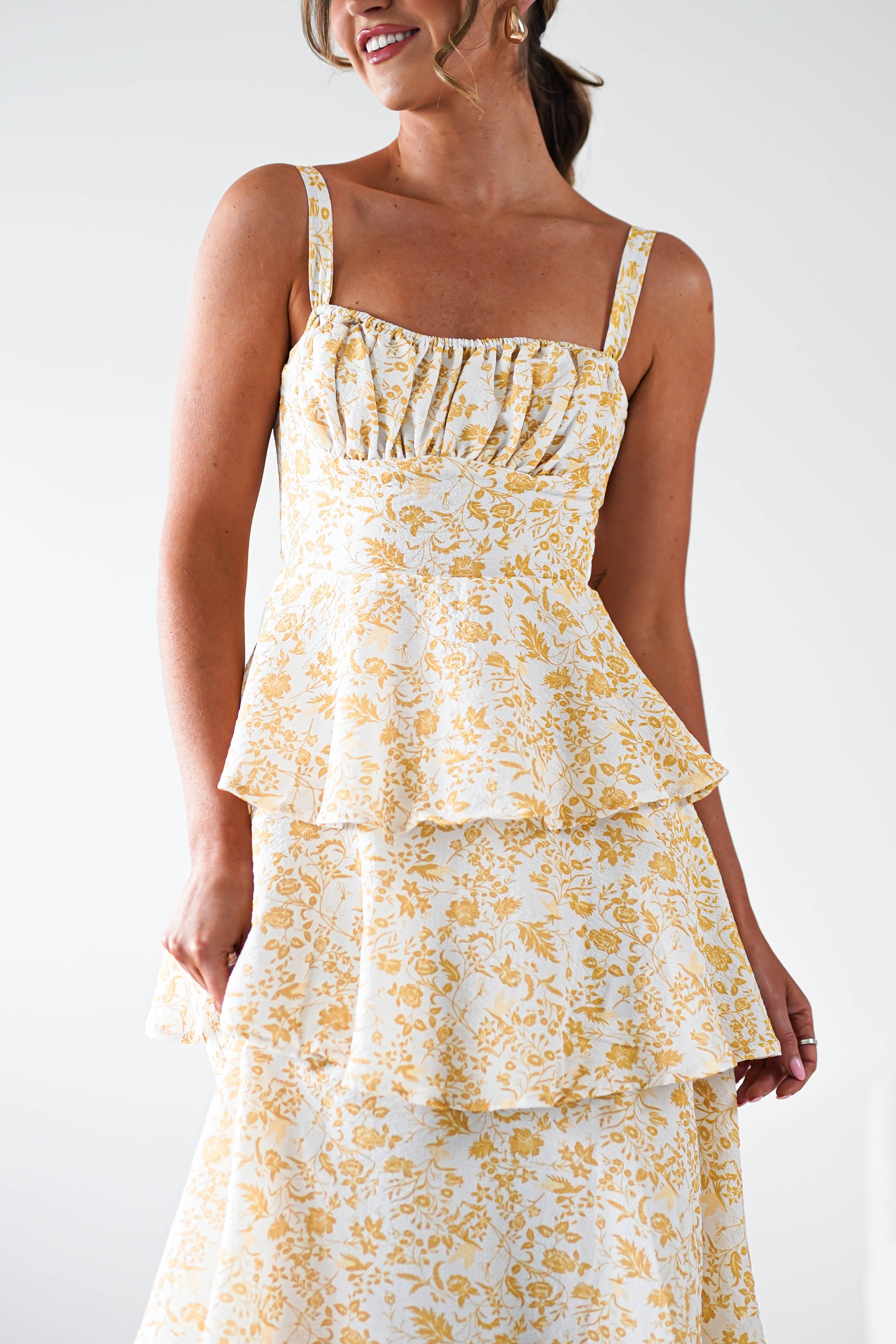 A woman wearing a yellow print maxi dress with ruffle detail and tiered layers stands smiling against a plain white background—perfect for summer events.
