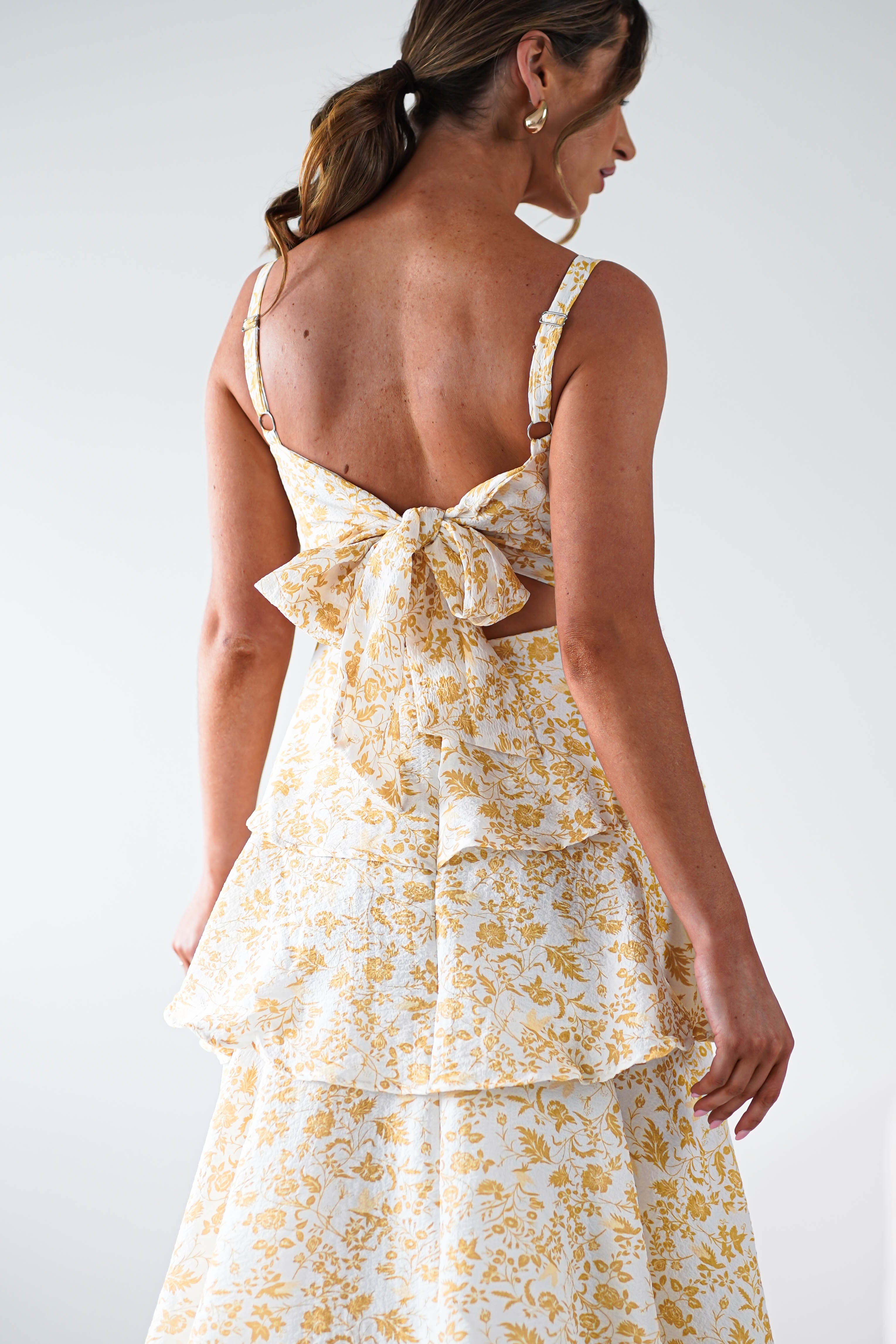 Woman wearing a sleeveless yellow print maxi dress with ruffle detail, a large bow at the back, and gold hoop earrings—perfect for summer events. She has brown hair tied in a ponytail and is facing away.