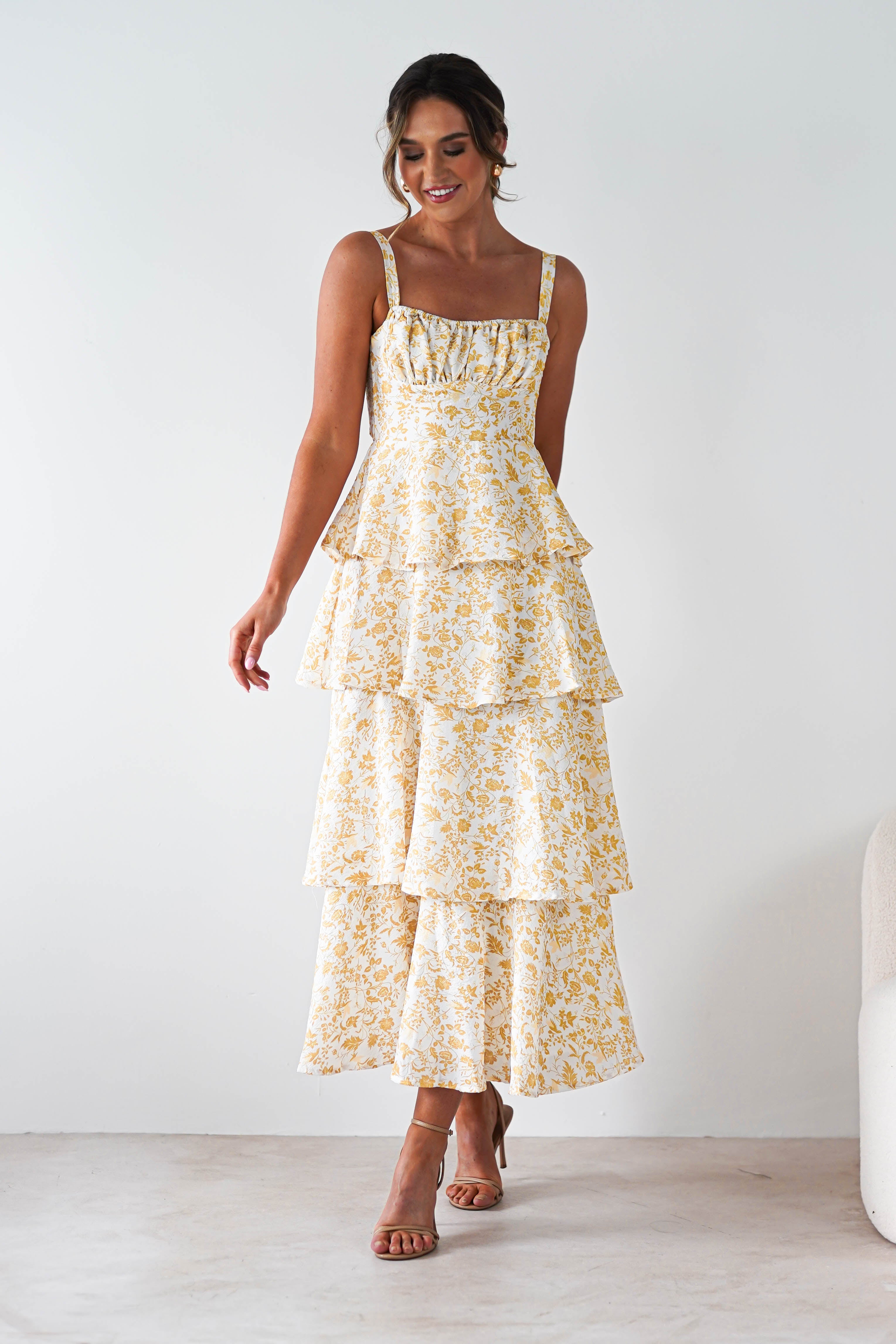 A woman wearing a yellow print maxi dress with ruffle detail and tiered layers stands smiling against a plain white background, paired with strappy heels—perfect for summer events.