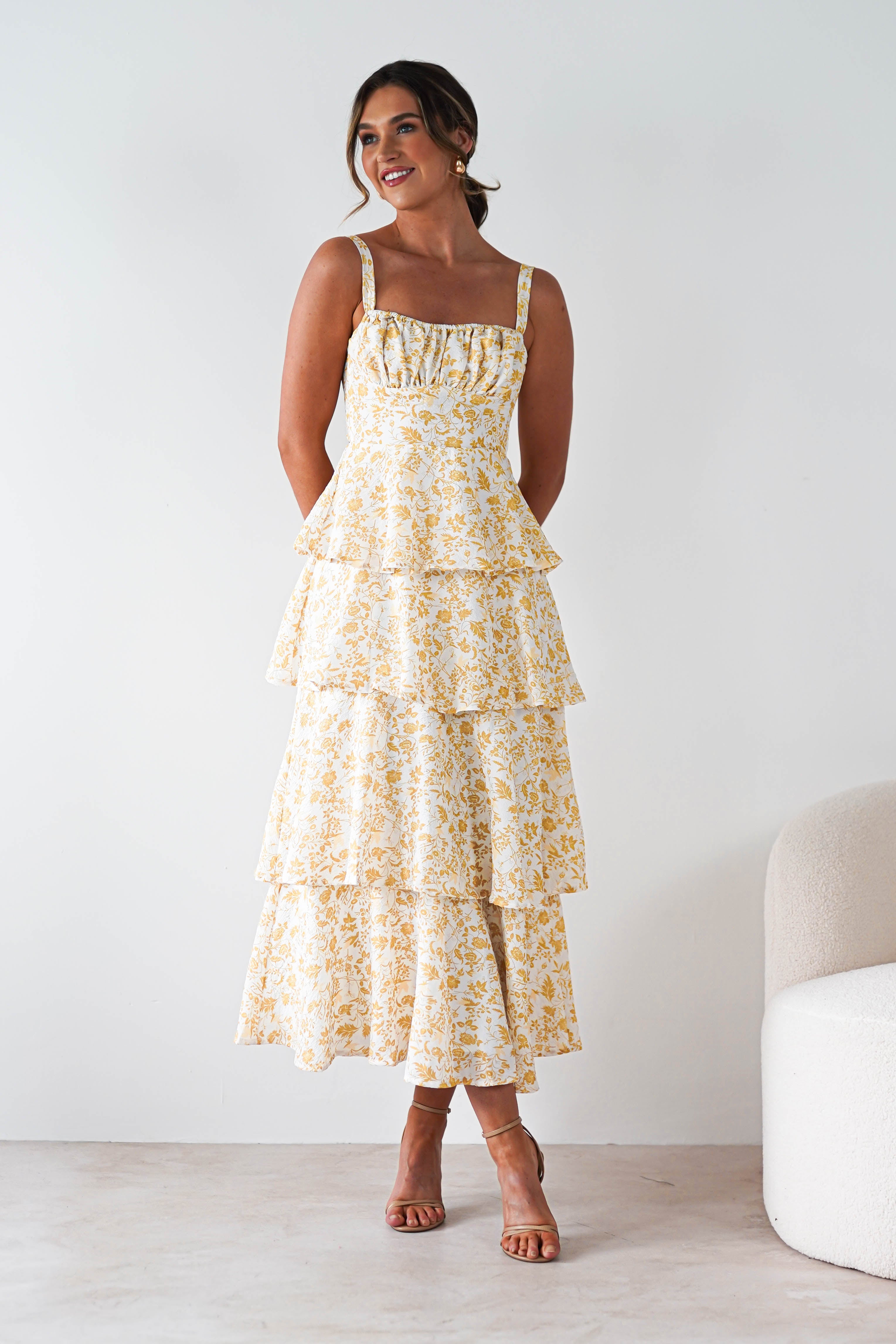 A woman stands indoors wearing a yellow print maxi dress with ruffle detail, perfect for summer events. She smiles with her hands behind her back, pairing the look with tan strappy heels. A light-colored chair is next to her.