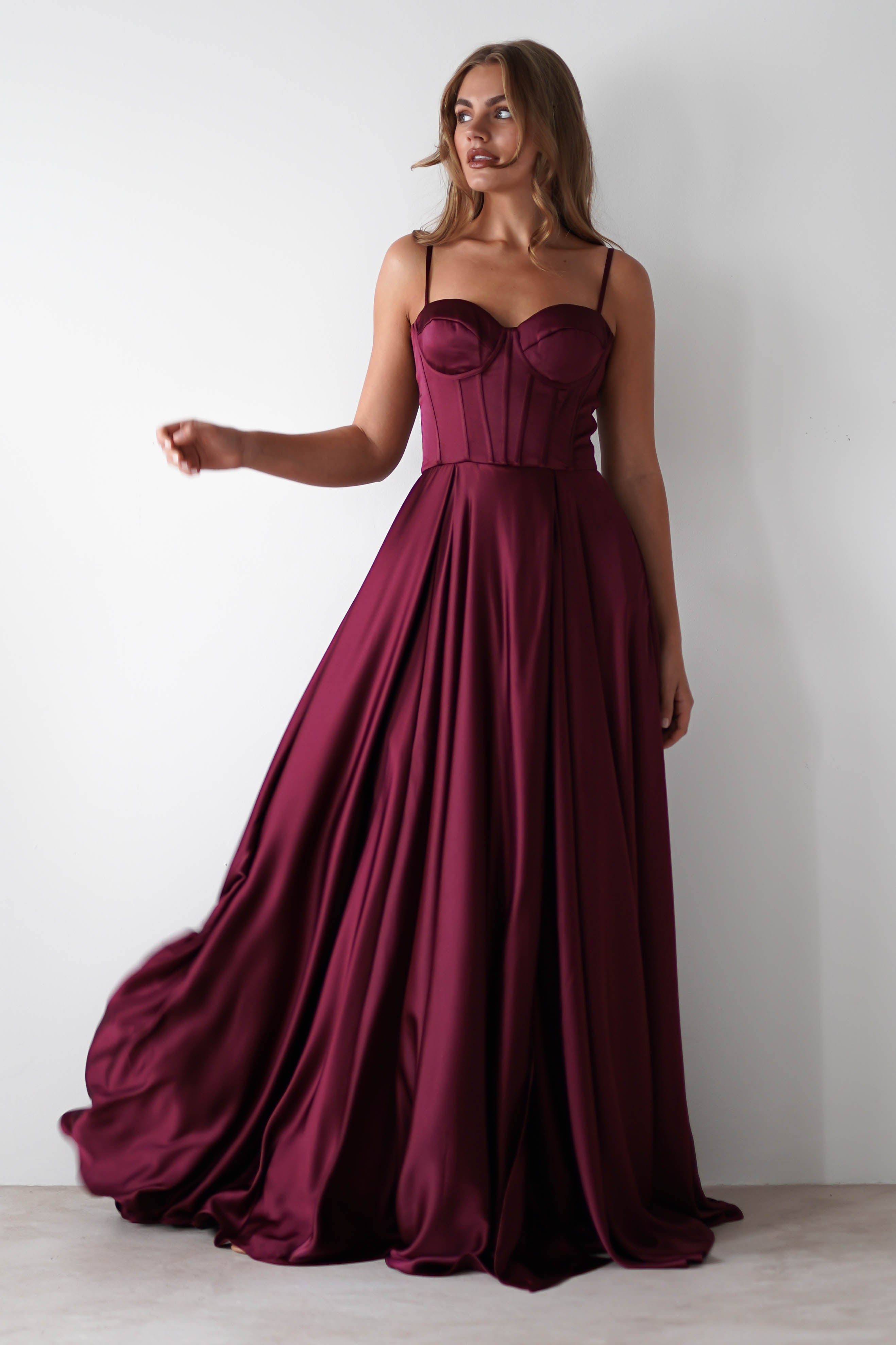 A woman stands against a plain white wall in a long, flowing burgundy satin gown with a lace-up back and fitted corset-style bodice—perfect for formal events. Her hair is loose, and she holds part of the dress, making it gently flare.