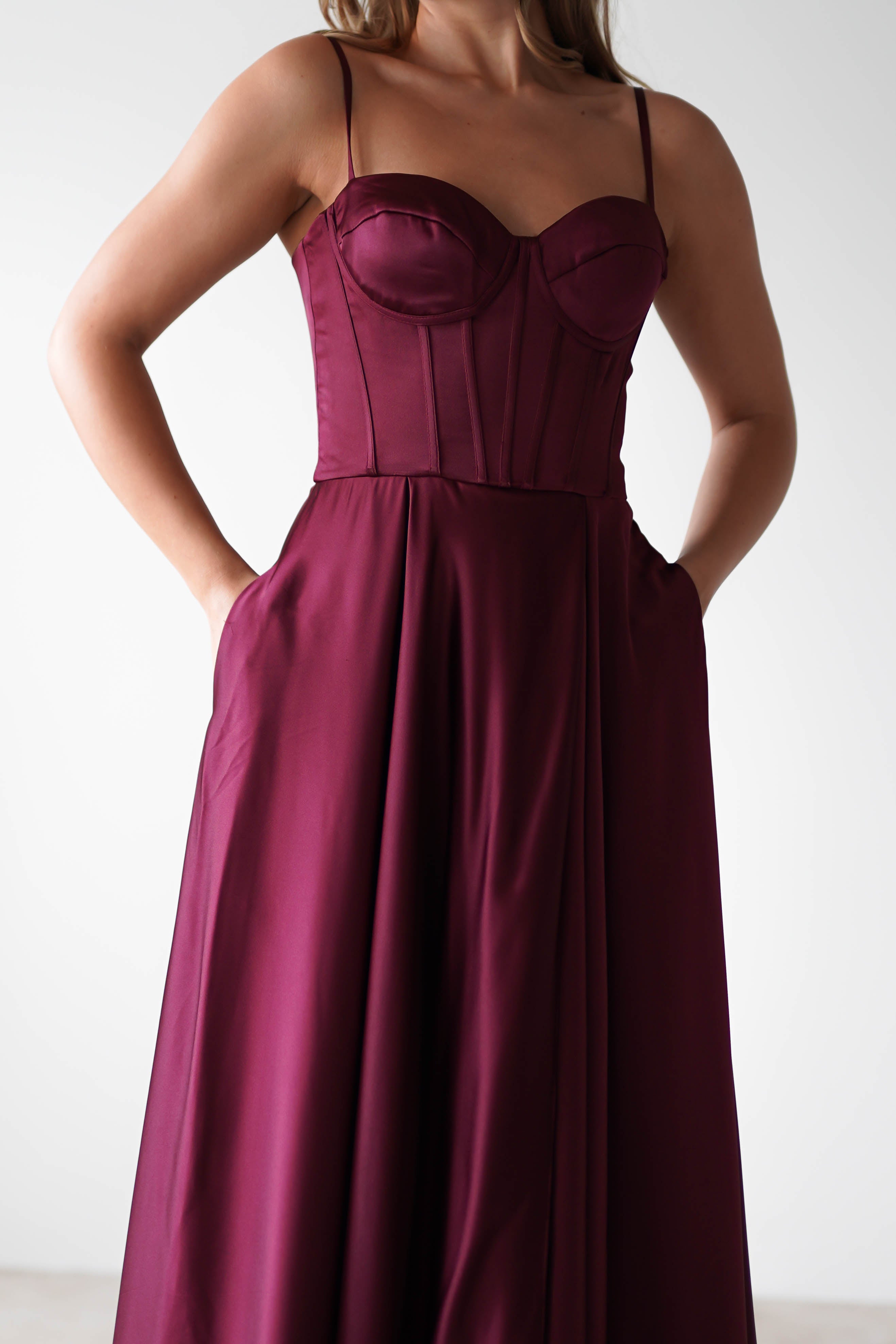 A woman wearing a sleeveless, burgundy satin gown with a fitted bodice, thin straps, lace-up back, and flared skirt stands with her hands in the dress pockets against a plain background—perfect for formal events.