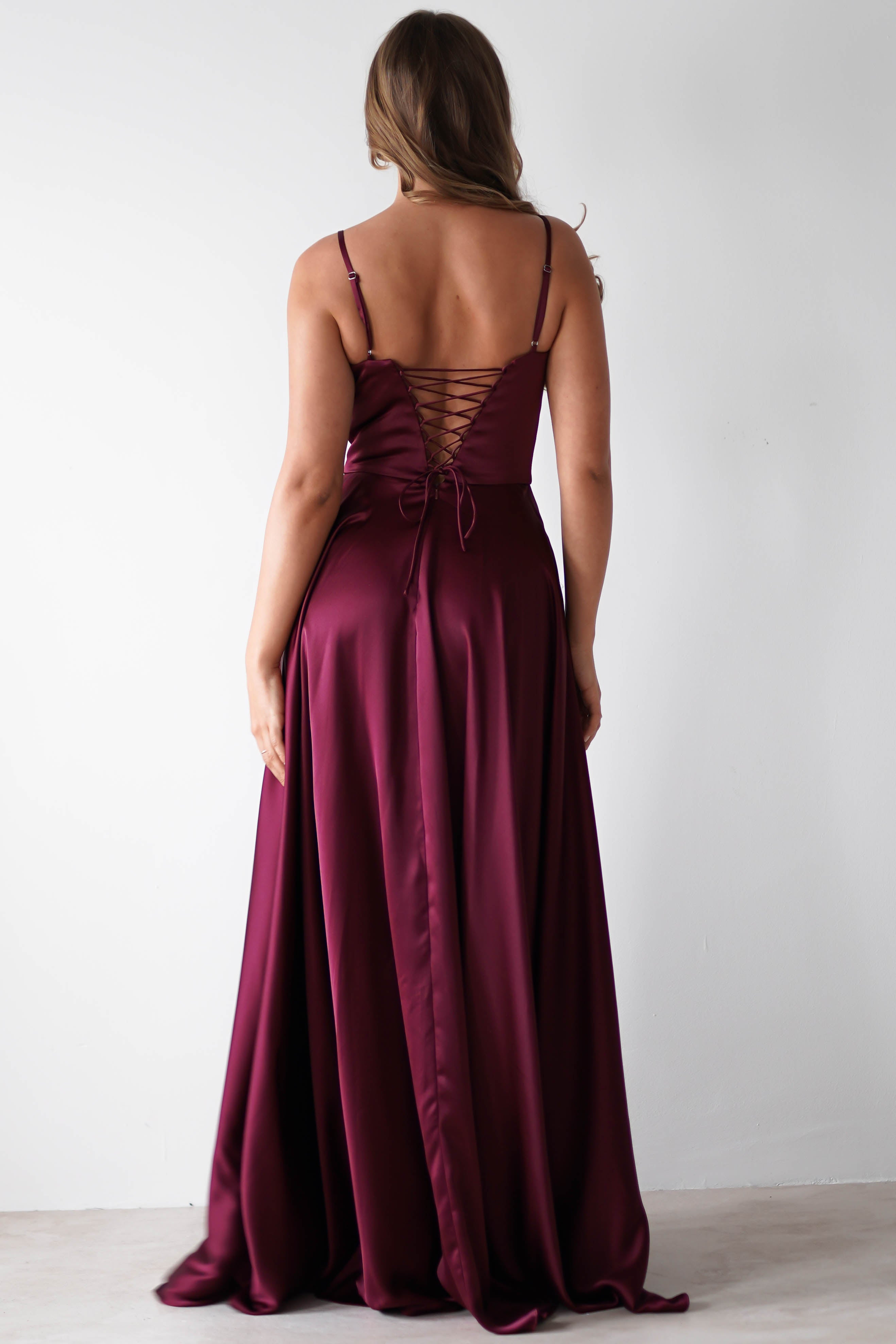 A woman with long, wavy hair wears a sleeveless satin gown with a lace-up back, standing facing away against a plain white background—a stunning choice for formal events.
