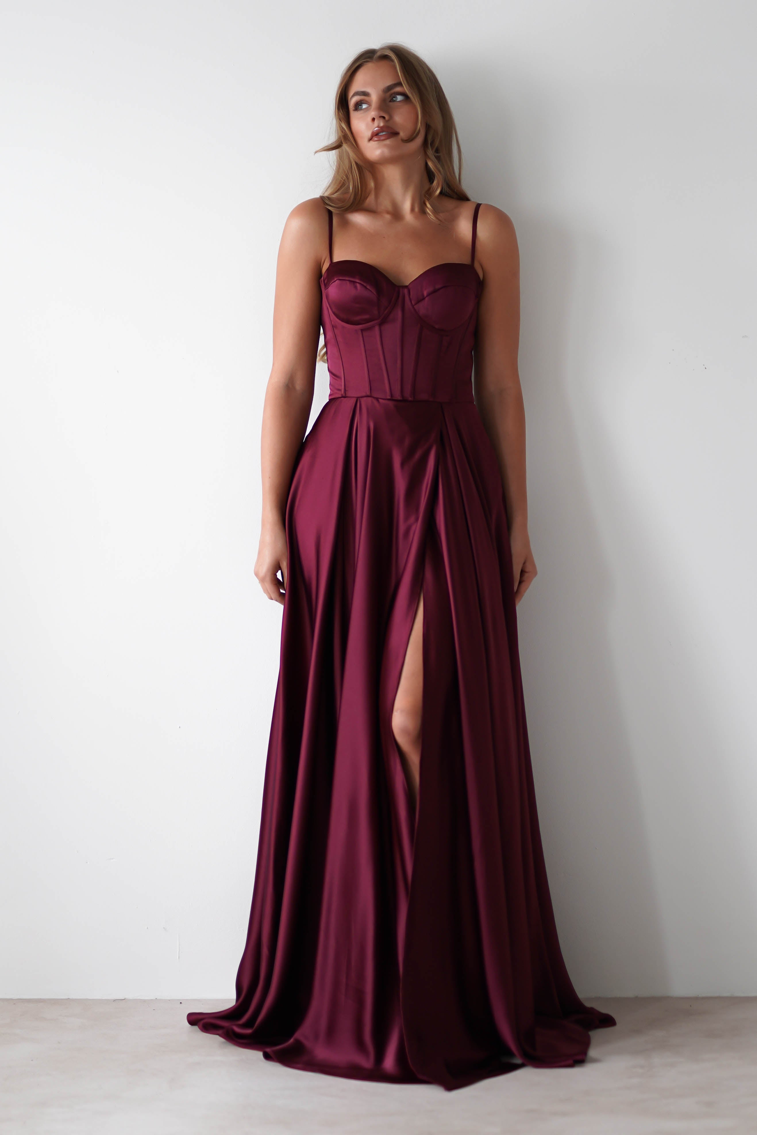 A woman wearing a sleeveless, burgundy satin gown with a lace-up back and thigh-high slit stands against a plain white wall, looking slightly upward—an elegant look perfect for formal events.