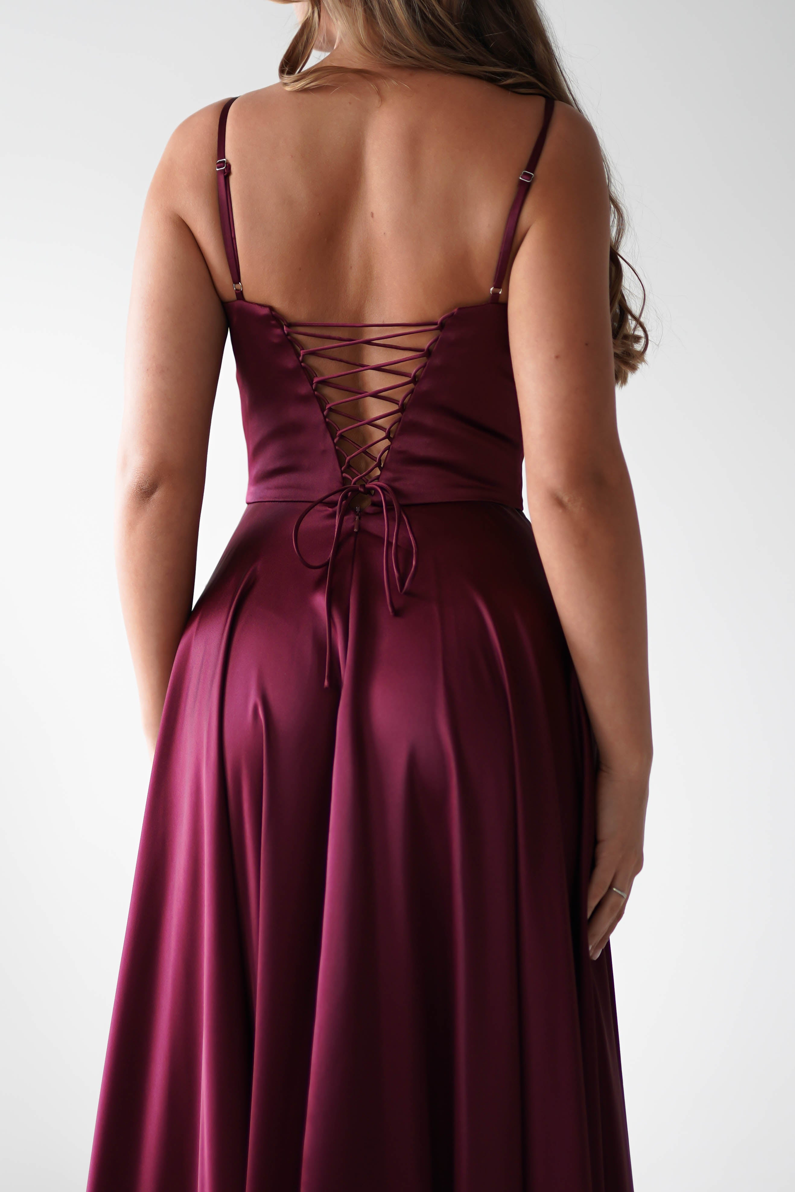 A woman wearing a sleeveless deep burgundy satin gown with a lace-up back detail stands facing away from the camera, her hair cascading over one shoulder—perfect for formal events.