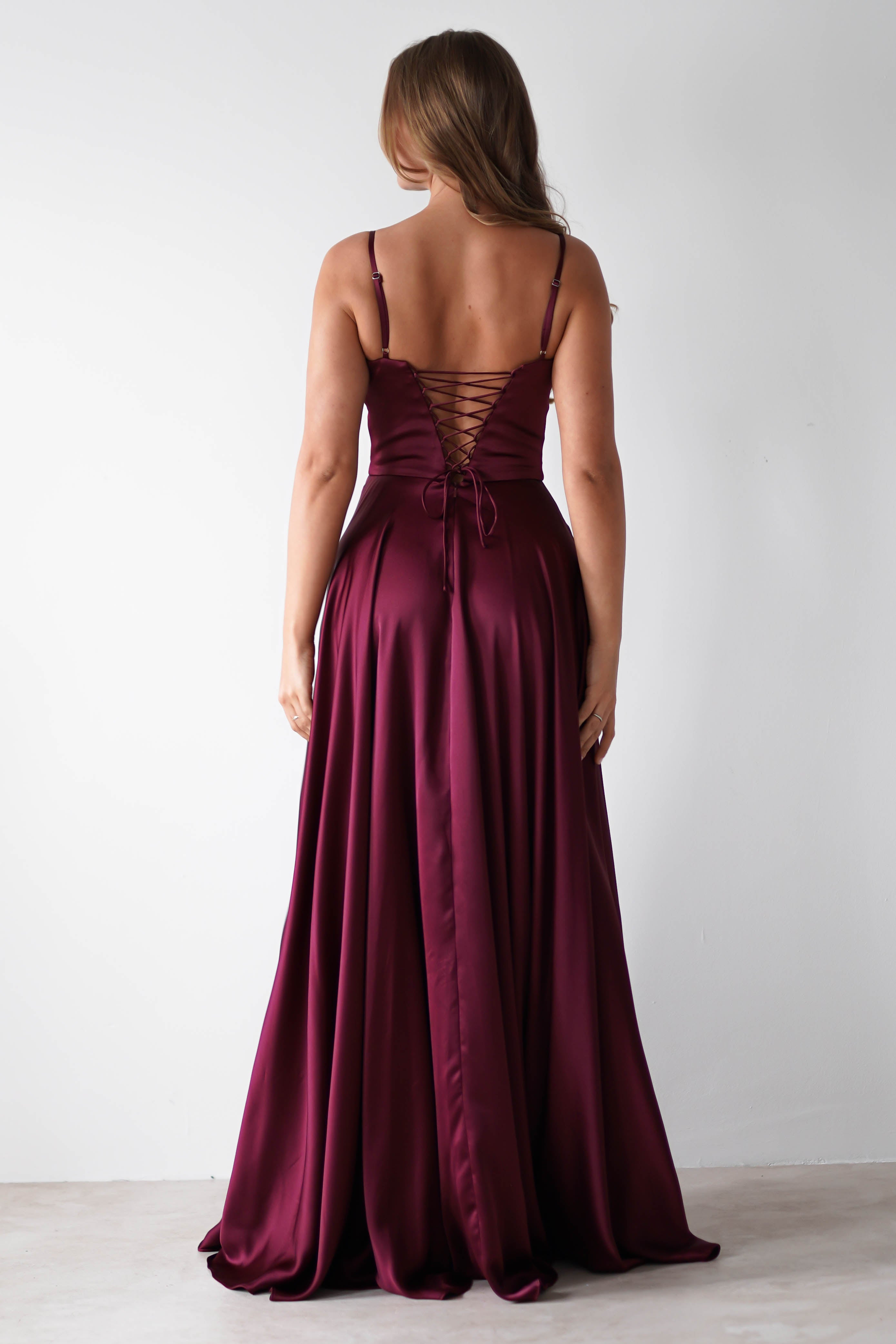 A woman stands facing away, wearing a satin gown with thin straps and a lace-up back—ideal for formal events. Her light brown hair is loose, and the background is a plain white wall.