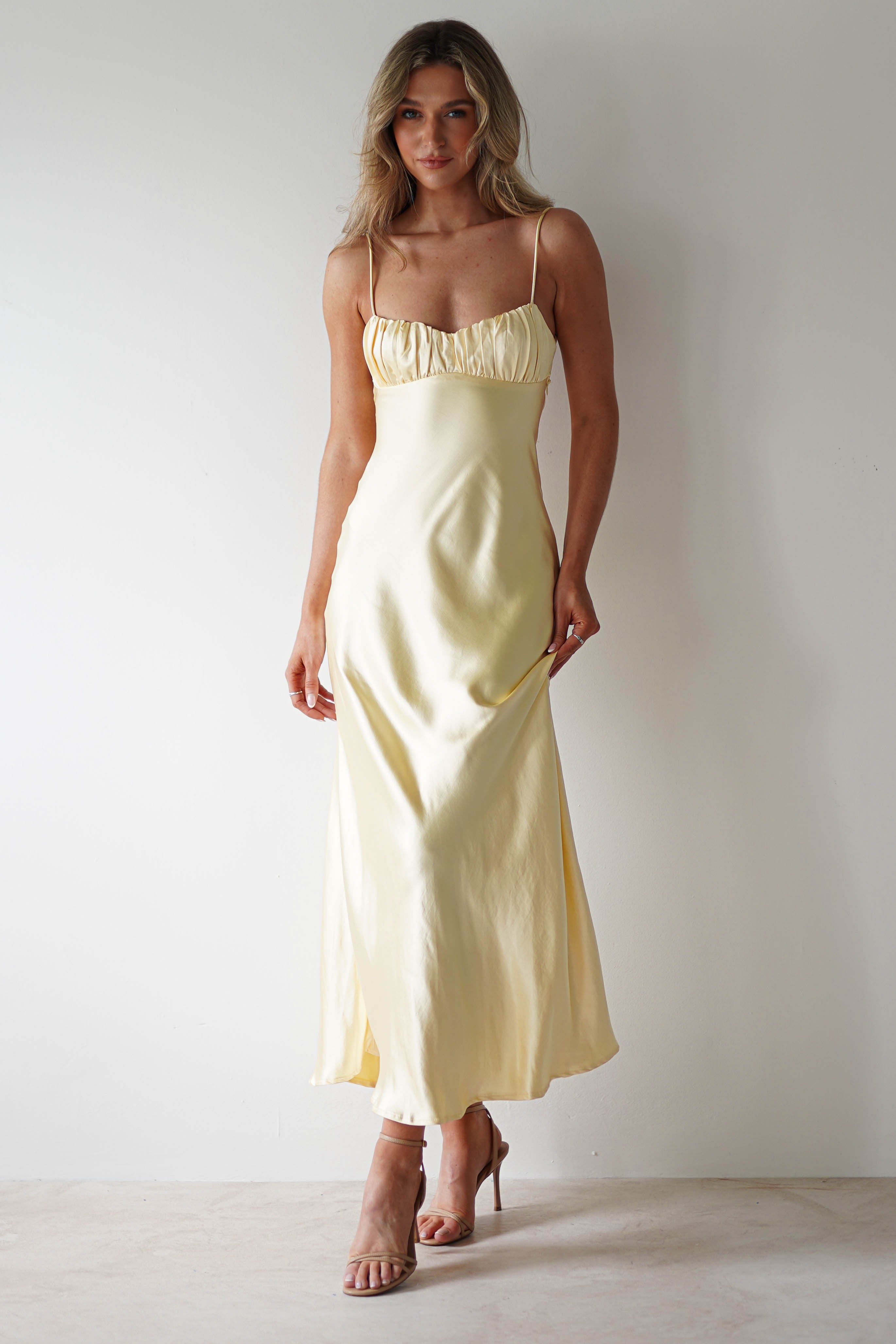 A woman wearing a pale yellow, satin spaghetti-strap midaxi dress with an open back and nude heels stands against a plain white background, looking forward with a slight smile.