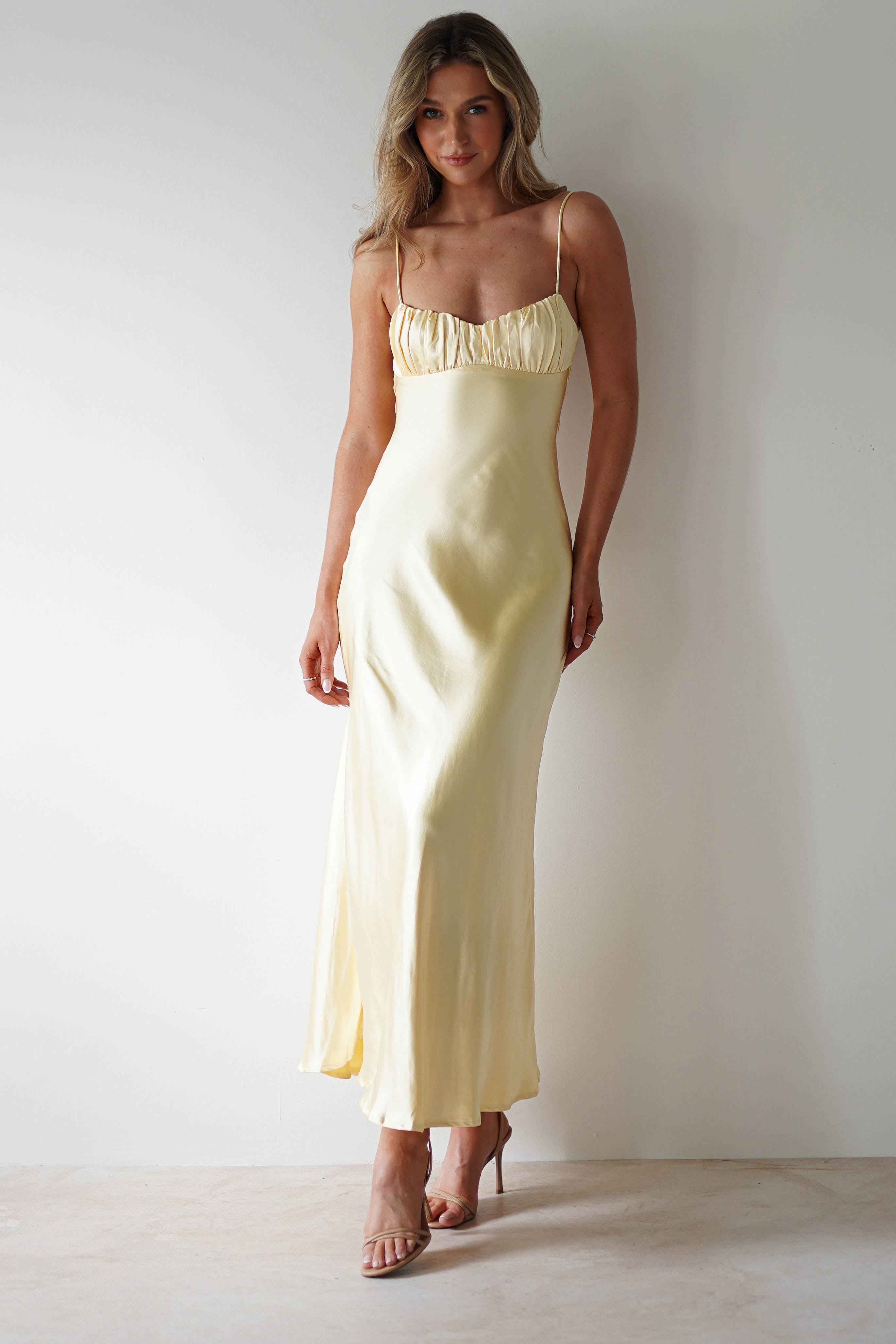 A woman stands against a plain white wall wearing a long, pale yellow satin midaxi dress with thin straps, an open back, and ruched detailing at the bust, paired with nude high-heeled sandals—perfect for a wedding guest look.