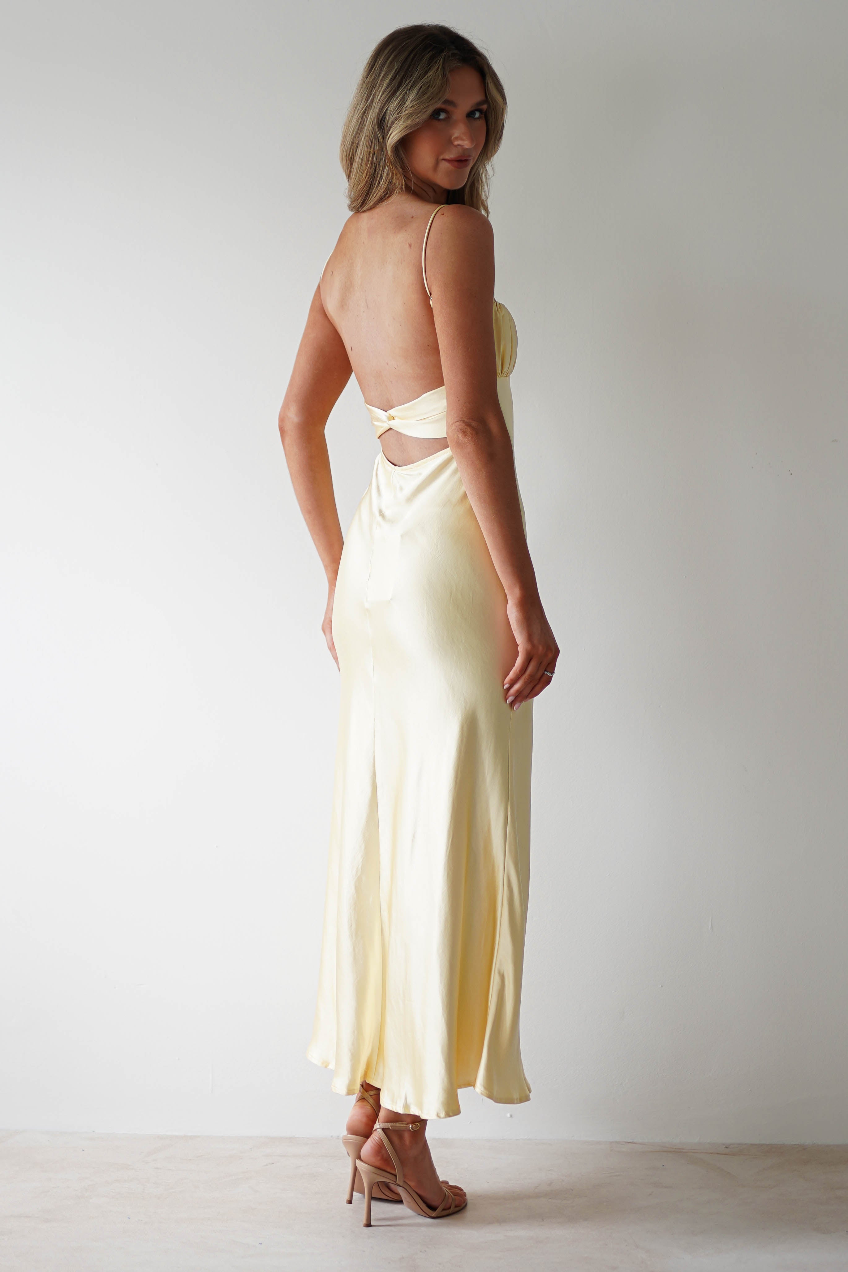 A woman with long, light brown hair poses in a pale yellow, open back satin midaxi dress with thin straps and strappy heels against a plain white background—an elegant look perfect for a wedding guest.