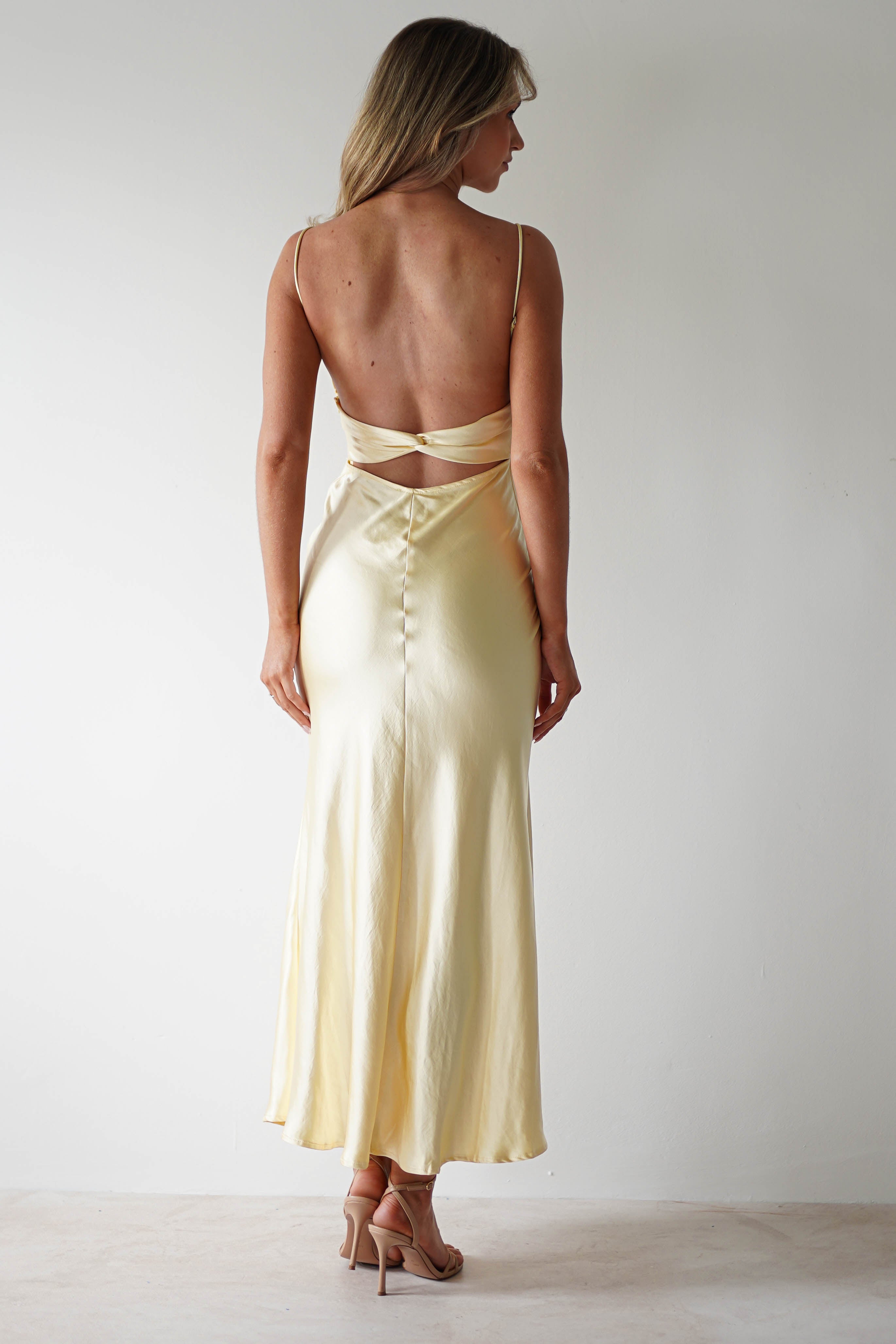 A woman stands facing away, wearing an open back, pale yellow satin midaxi dress with thin straps and button details down the skirt, paired with nude heels, against a plain white background. Perfect for a stylish wedding guest look.