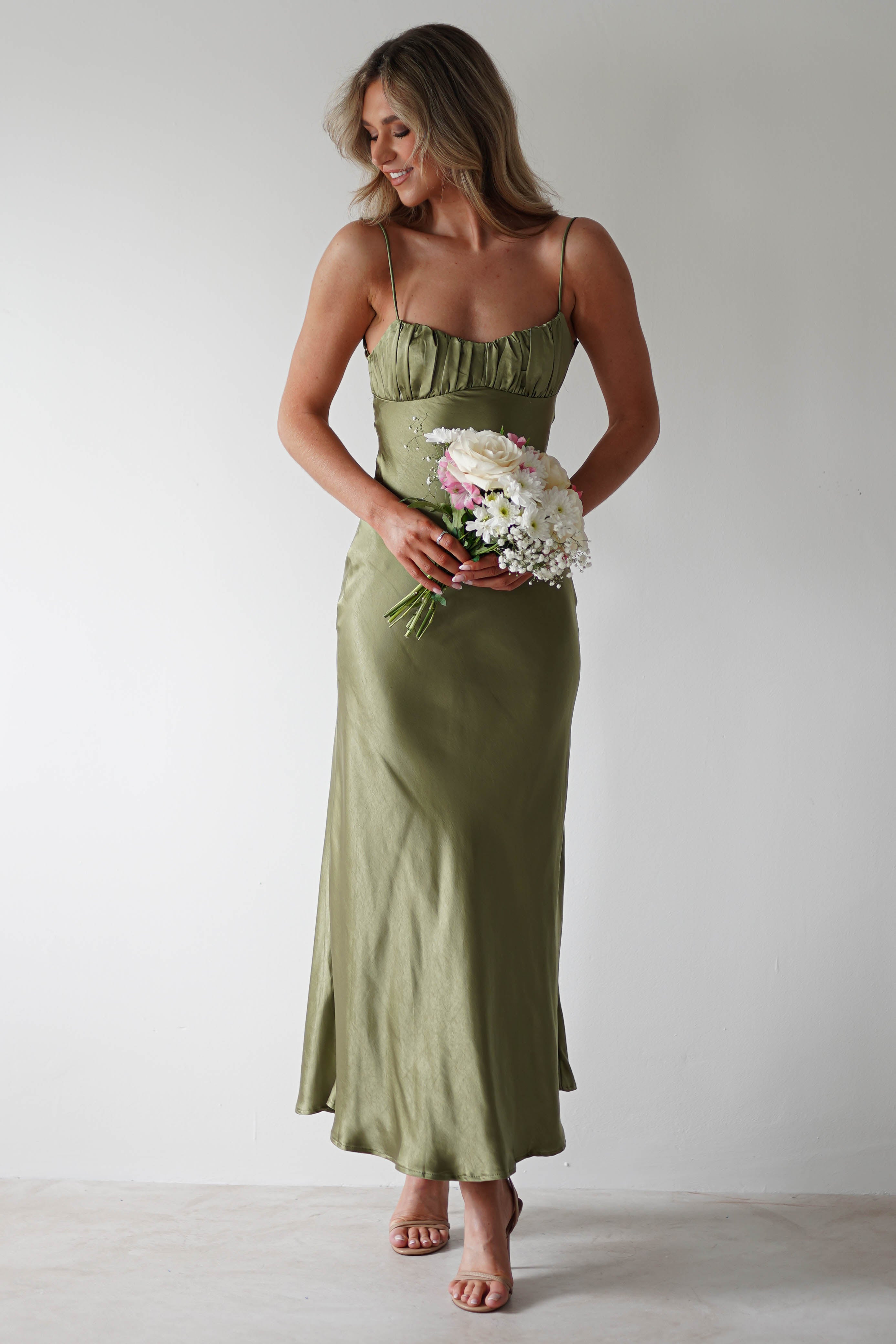 A woman in a light olive green satin midaxi dress holds a bouquet of white and pink flowers, standing against a plain white background, smiling and looking to the side—perfect as a stylish wedding guest look.