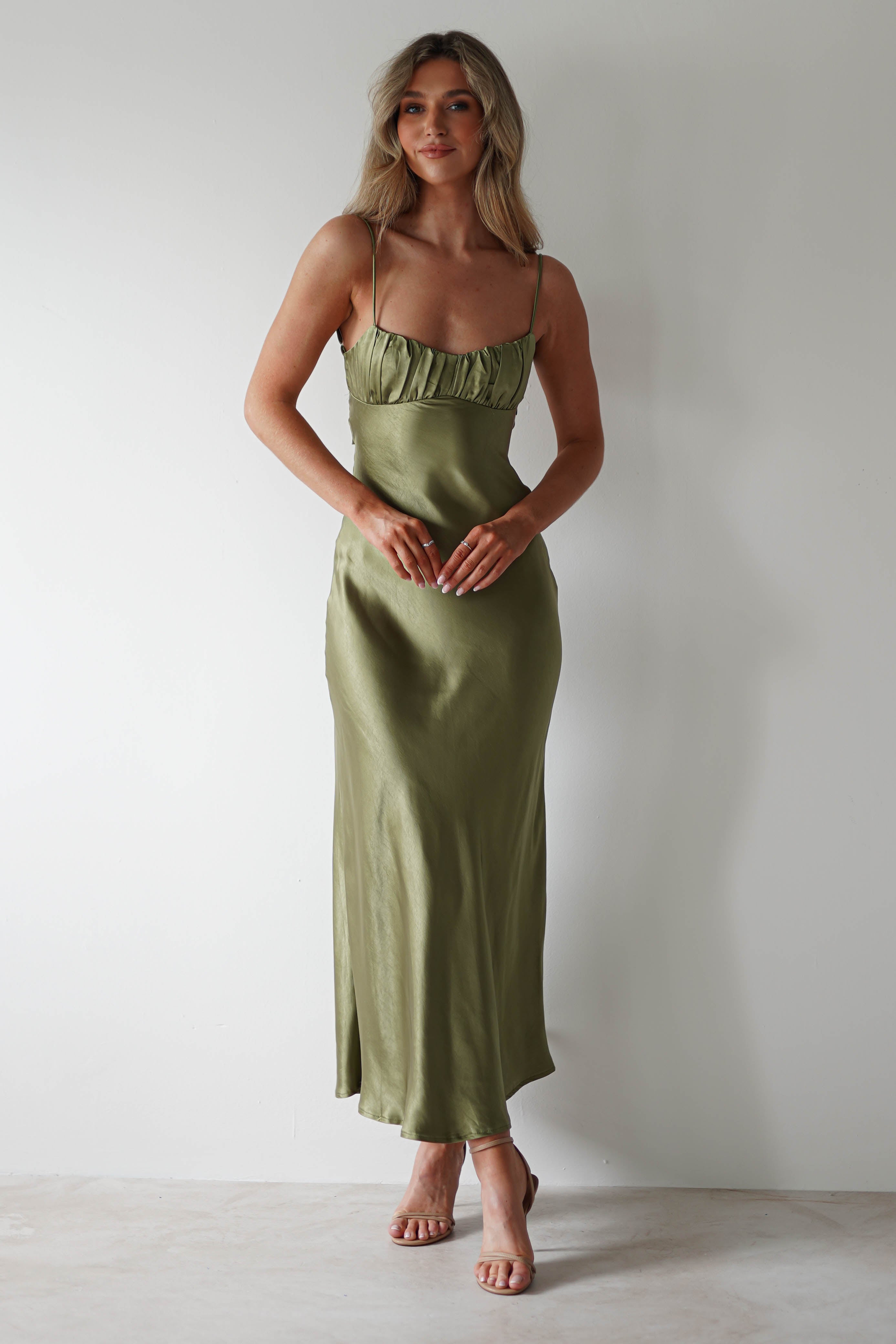 A woman stands against a plain white wall, wearing a long, olive green satin midaxi dress with thin straps and ruffled detailing on the bodice—an elegant choice for a wedding guest. She has long, wavy blonde hair and nude heels.