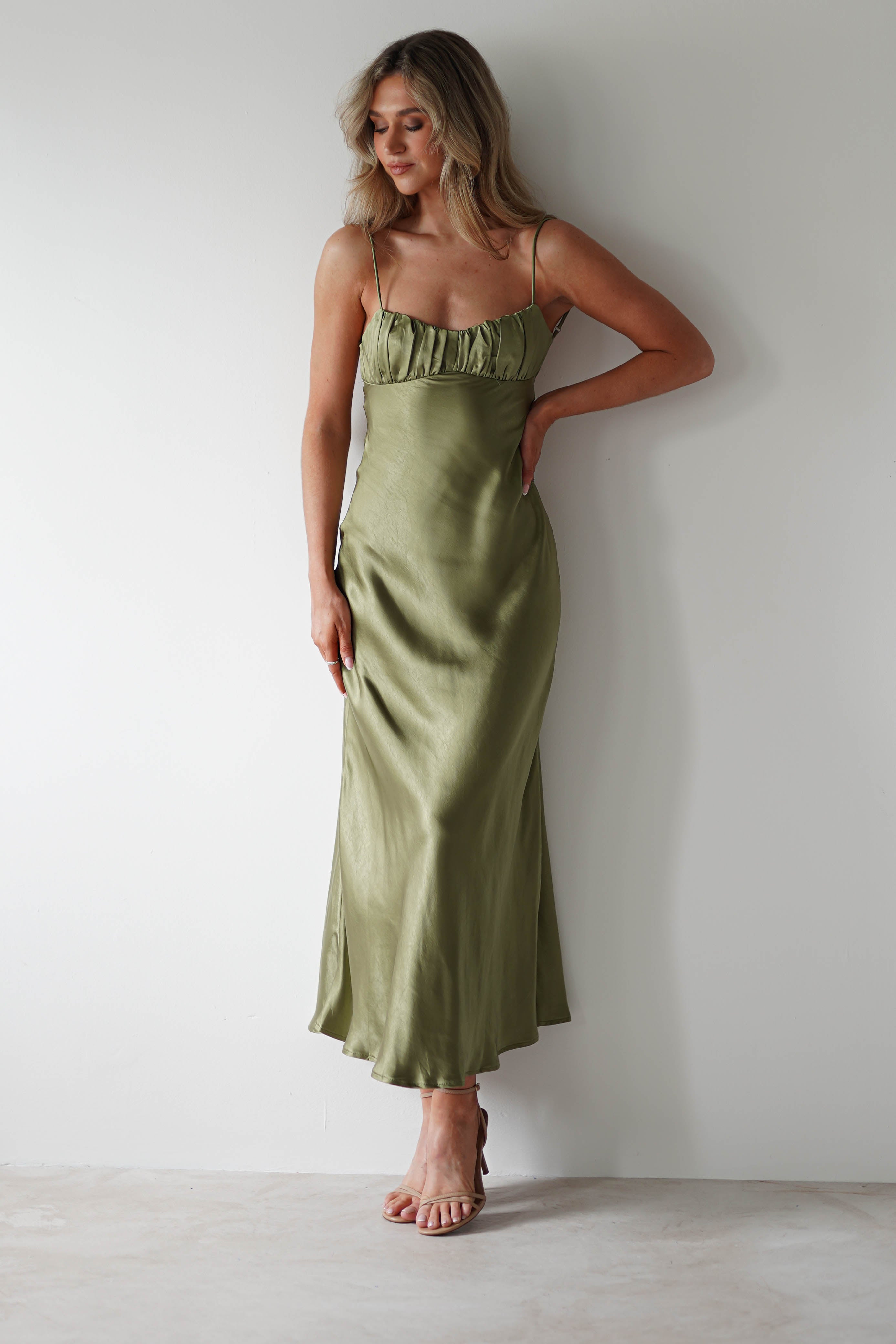 A woman in a light olive green polyester midaxi dress with thin straps stands against a plain white wall, her left hand on her hip and right hand relaxed by her side. She wears strappy sandals, perfect for a wedding guest, and smiles softly while looking downward.