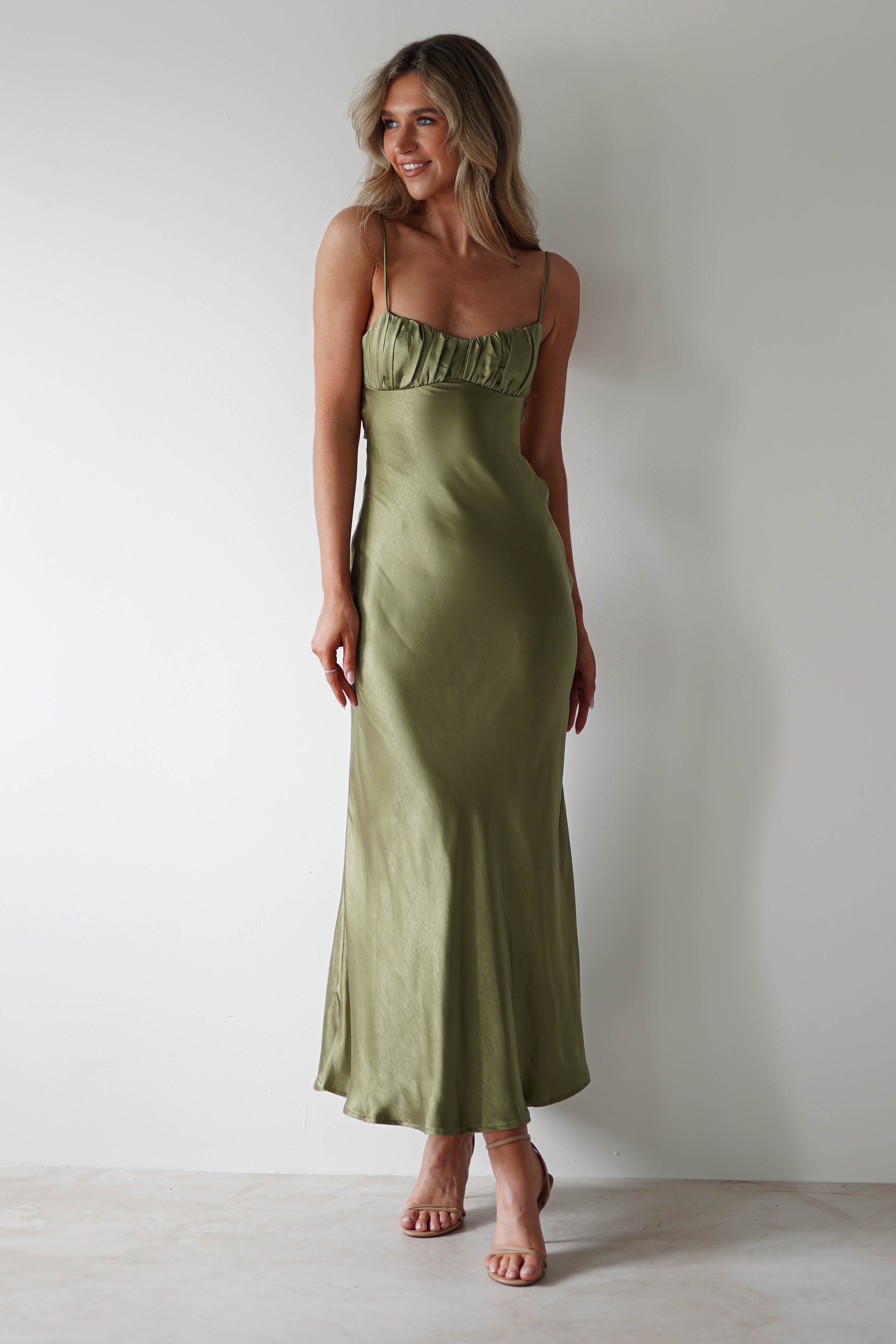 A woman wearing a light olive green, polyester midaxi dress with thin straps and ruffle detail at the bust stands against a plain white wall, smiling and looking to the side—perfect as a stylish wedding guest look.