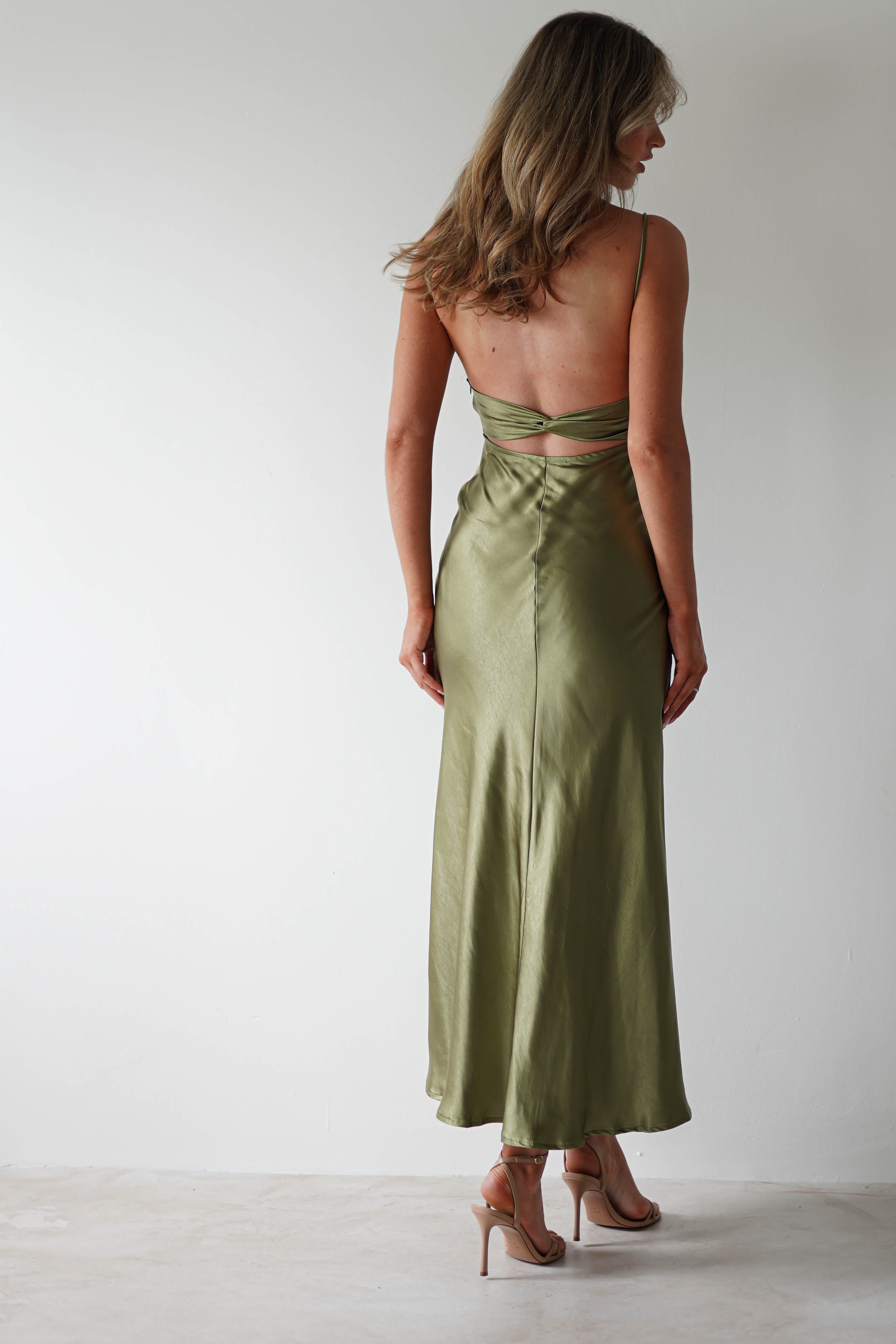 A woman with wavy, shoulder-length hair wears a shiny, olive green polyester midaxi dress and nude high heels, standing against a plain white background and facing away from the camera—perfect for a stylish wedding guest look.