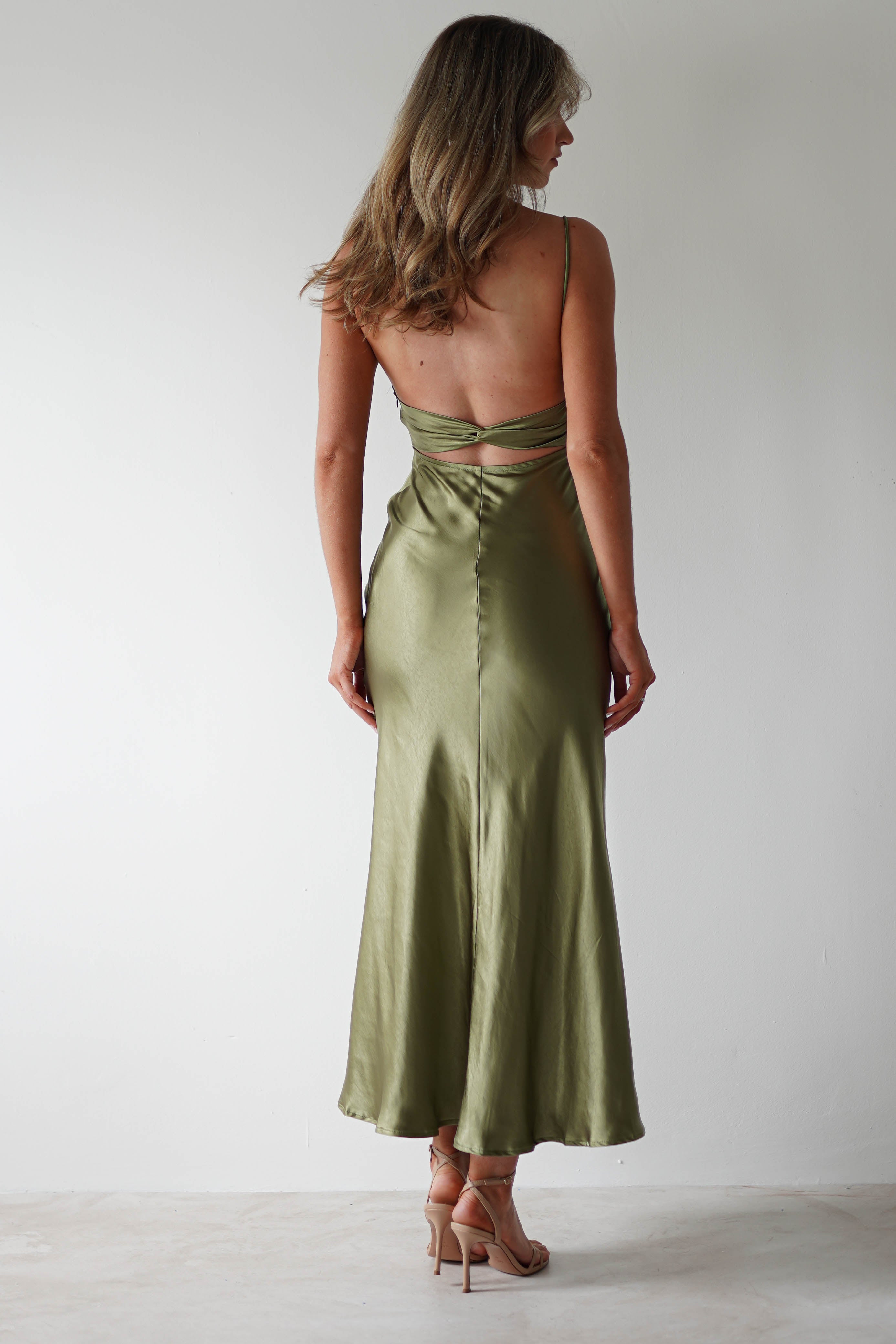 A woman stands facing away, wearing a backless, olive green polyester midaxi dress with thin straps and nude heels, against a plain white background. Her hair is loose and wavy, falling elegantly over her shoulders—a stunning look for any wedding guest.