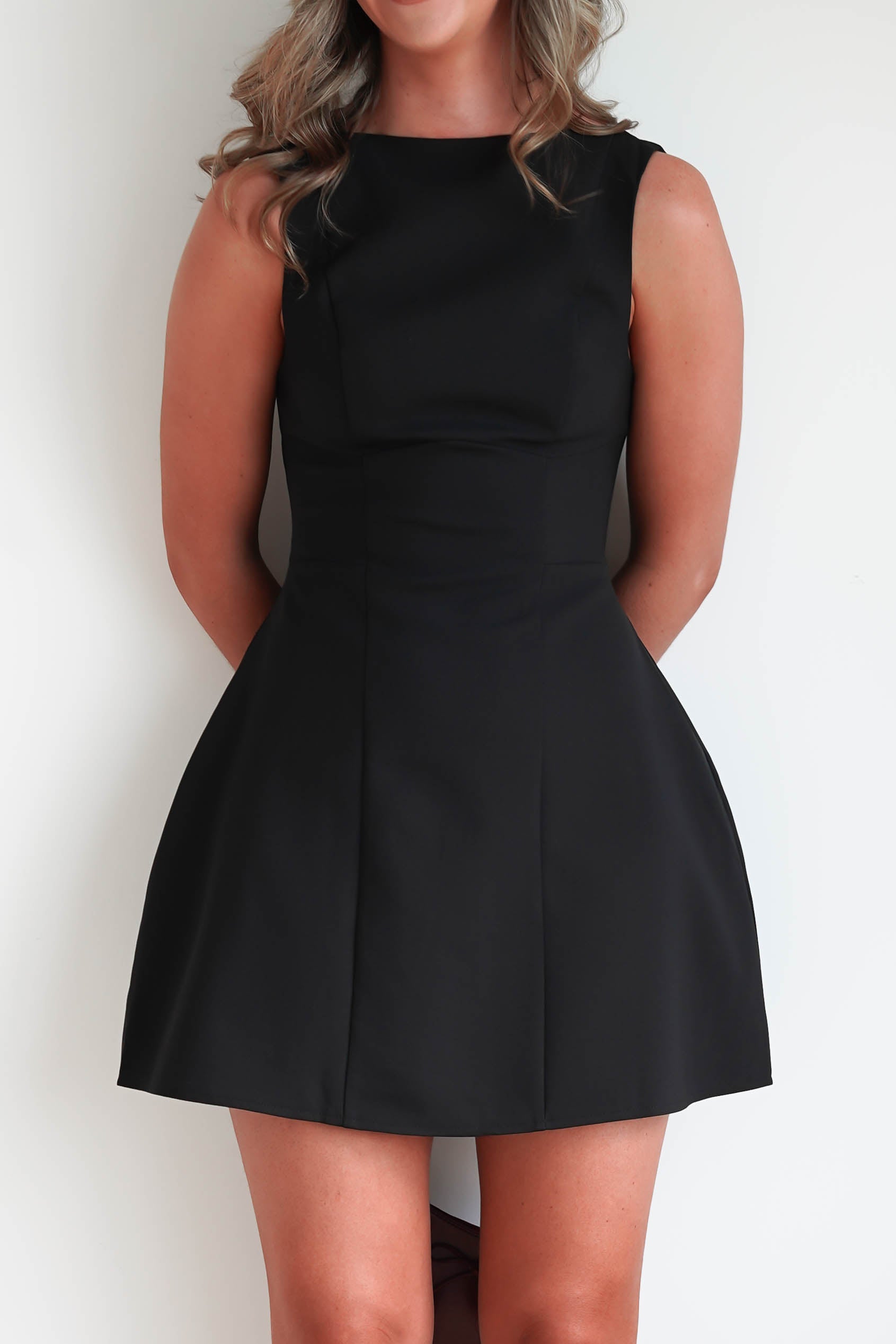 A woman in a sleeveless black mini dress stands against a plain white background. Perfect for graduations, the dress is fitted at the top and flares out into a short, A-line skirt. Her arms are positioned behind her back, and her head is cropped out of the frame.