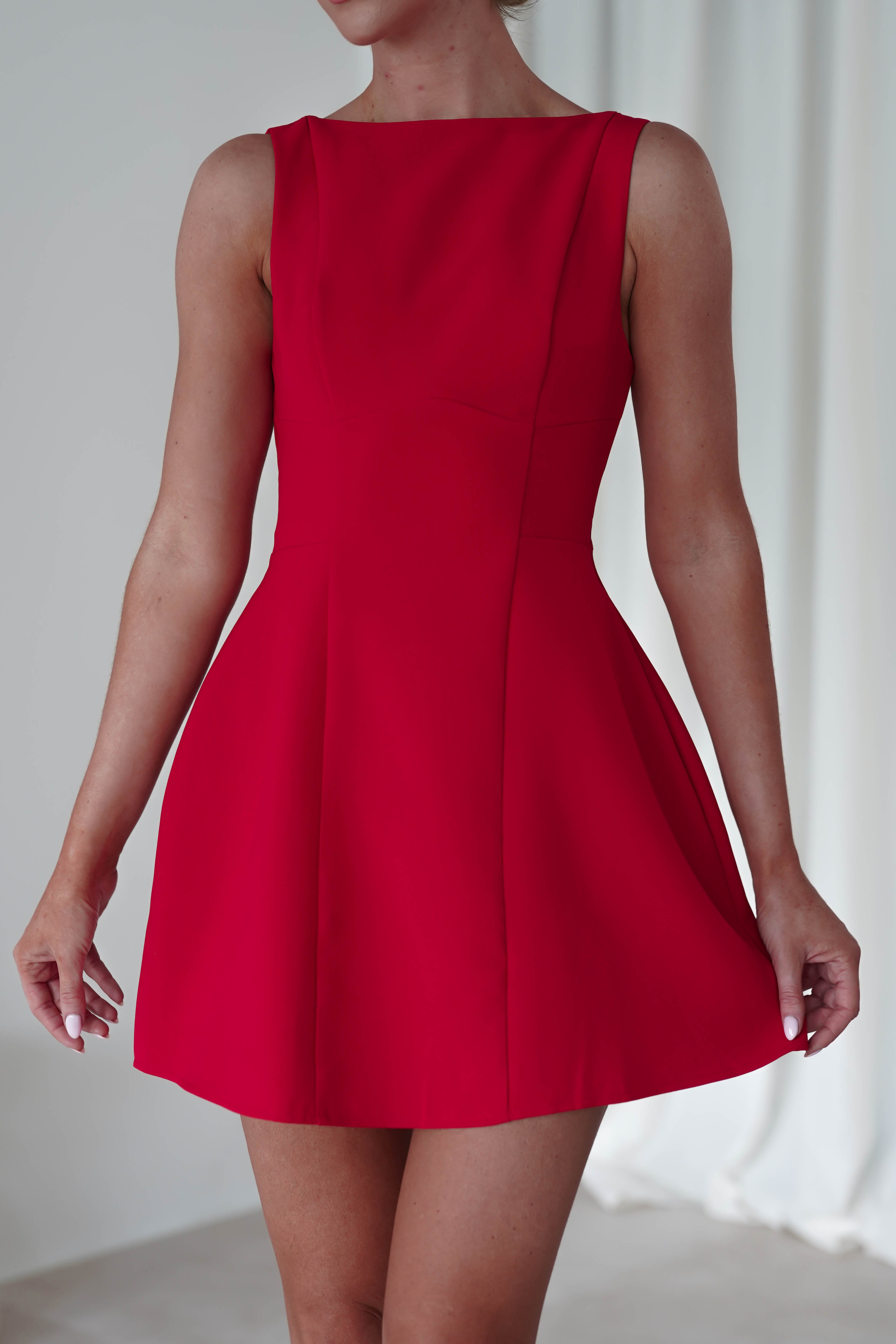 A person is wearing a sleeveless, bright red party dress. The image focuses on the dress and the persons arms and upper body, with a blurred neutral background. The wedding guest dress features a fitted bodice and flares out towards the hem.