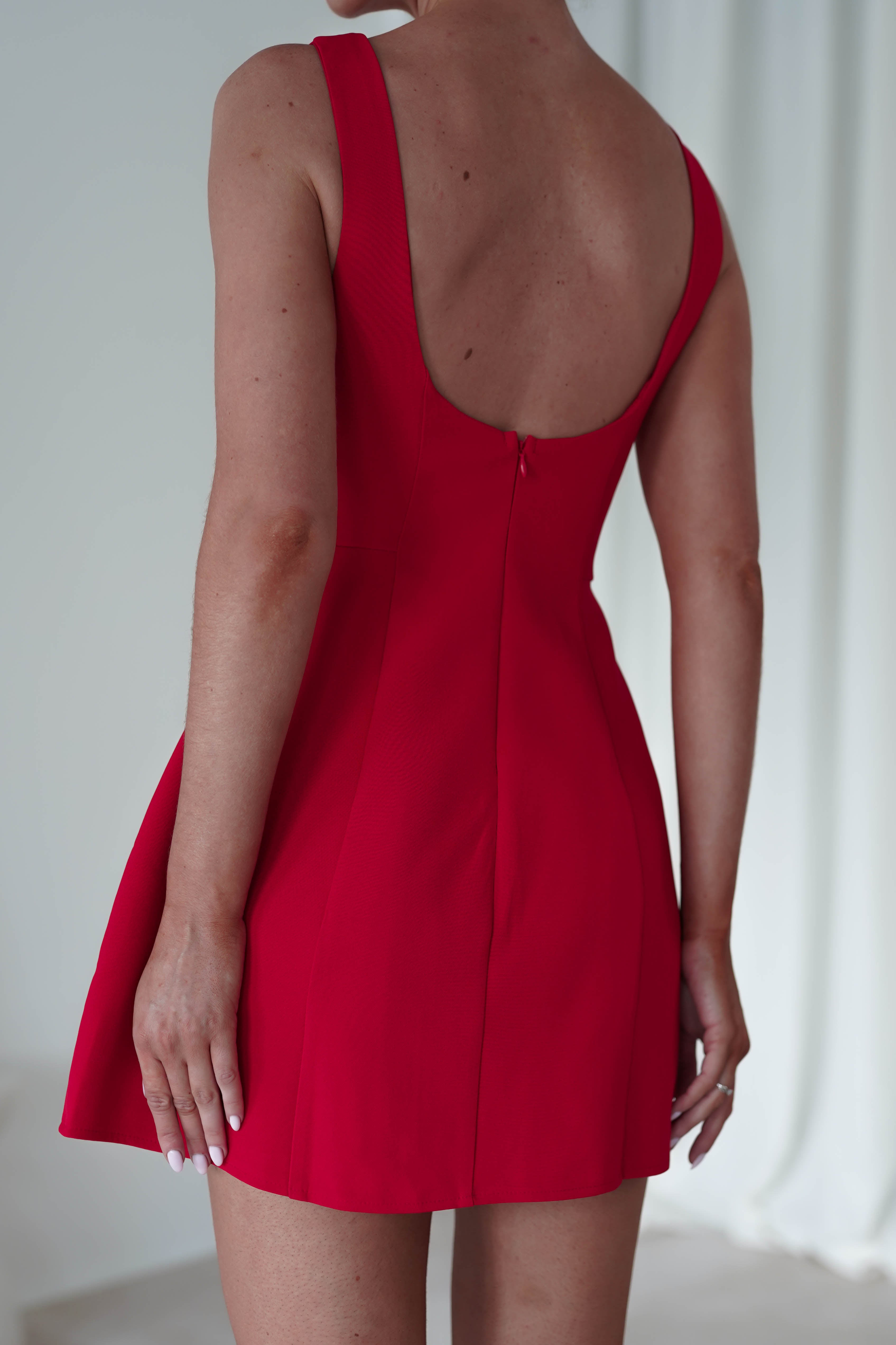 A person wearing a sleeveless red party dress is standing with their back to the camera. The dress features a low scoop back and a flared skirt. The background is softly blurred, giving a neutral tone to the setting.