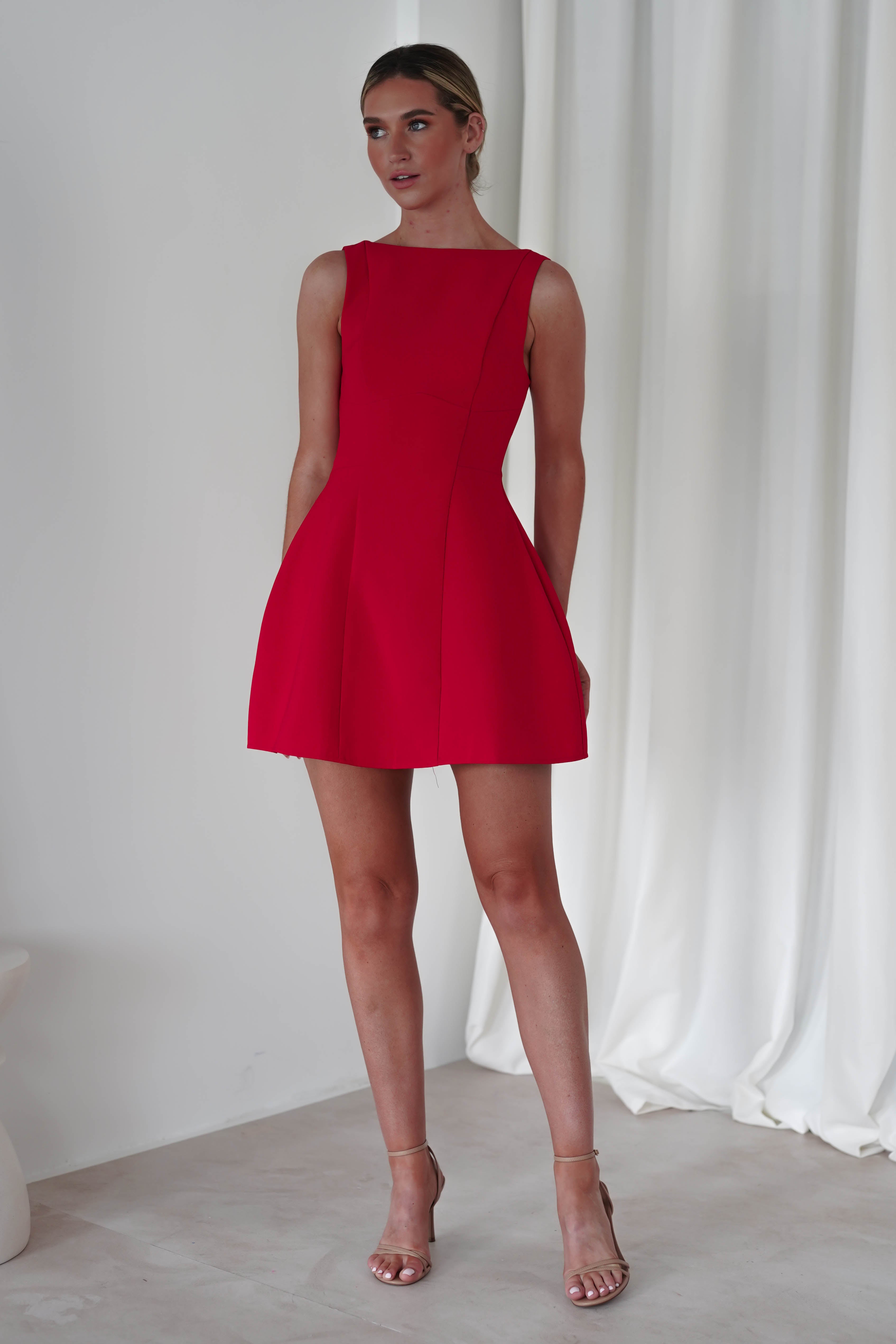 A person stands against a white curtain, wearing a sleeveless, fitted red mini dress with a flared skirt. They have short hair styled back and are wearing strappy high-heeled sandals—a perfect choice for a party or as a wedding guest dress.