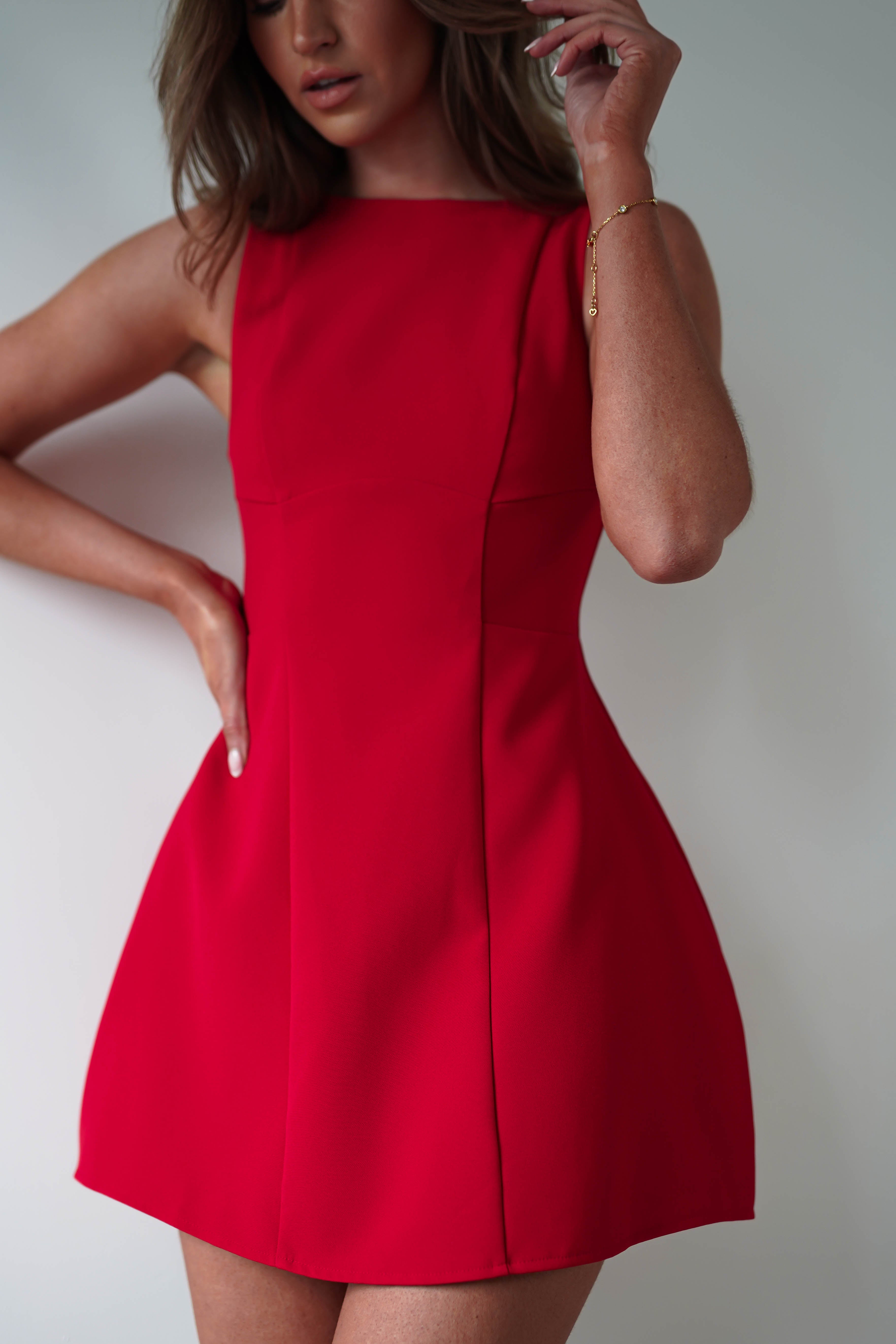 A person in a sleeveless, fitted red mini dress stands against a plain background, slightly cropped from the neck up. One hand rests on their hip while the other adjusts their hair, perfectly embodying the essence of a chic party dress.