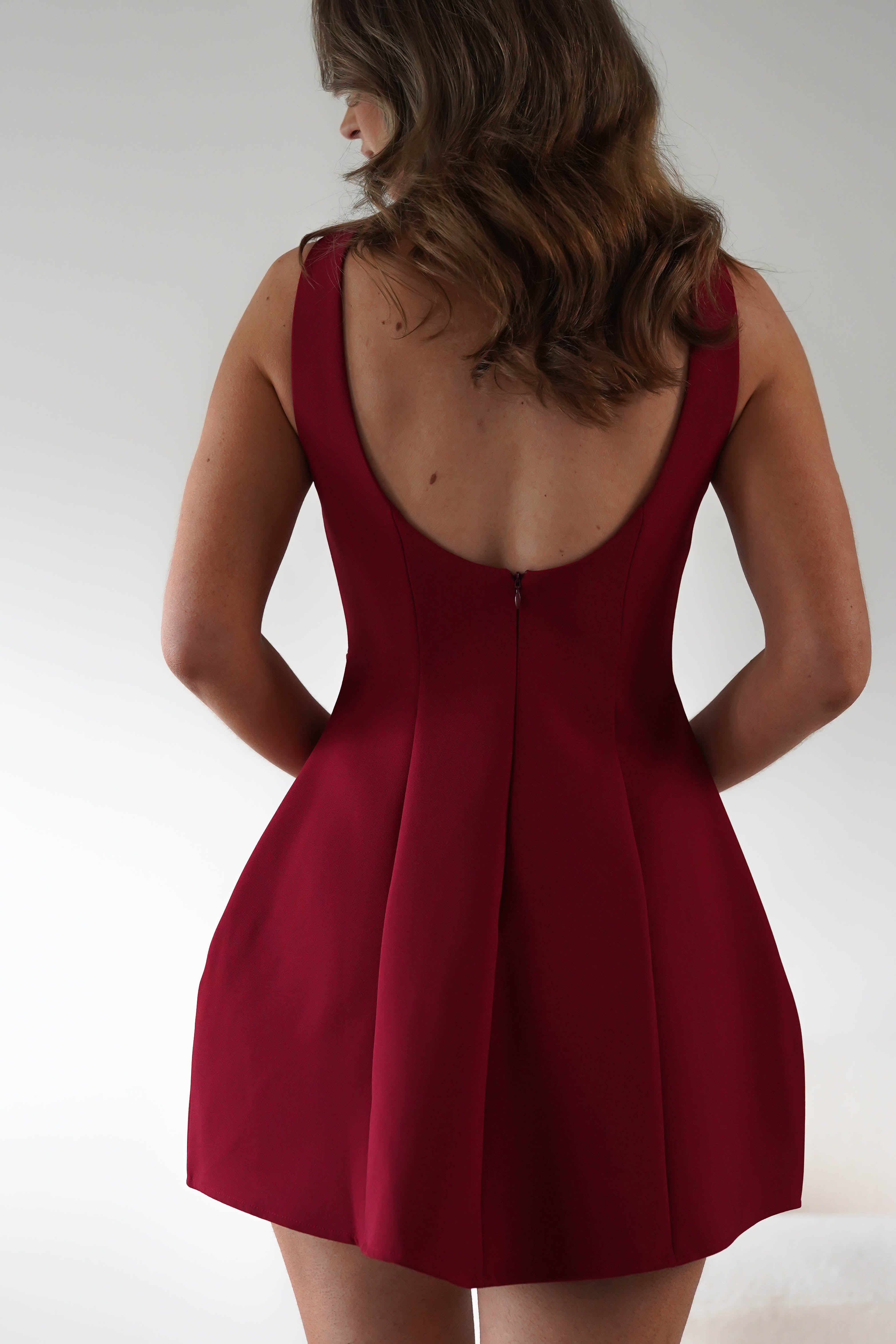 A woman with wavy brown hair stands with her back to the camera, wearing a sleeveless, deep red dress featuring a low back and pleated skirt—perfect for a classic cut look at graduations or as a wedding guest. The background is plain and softly lit.