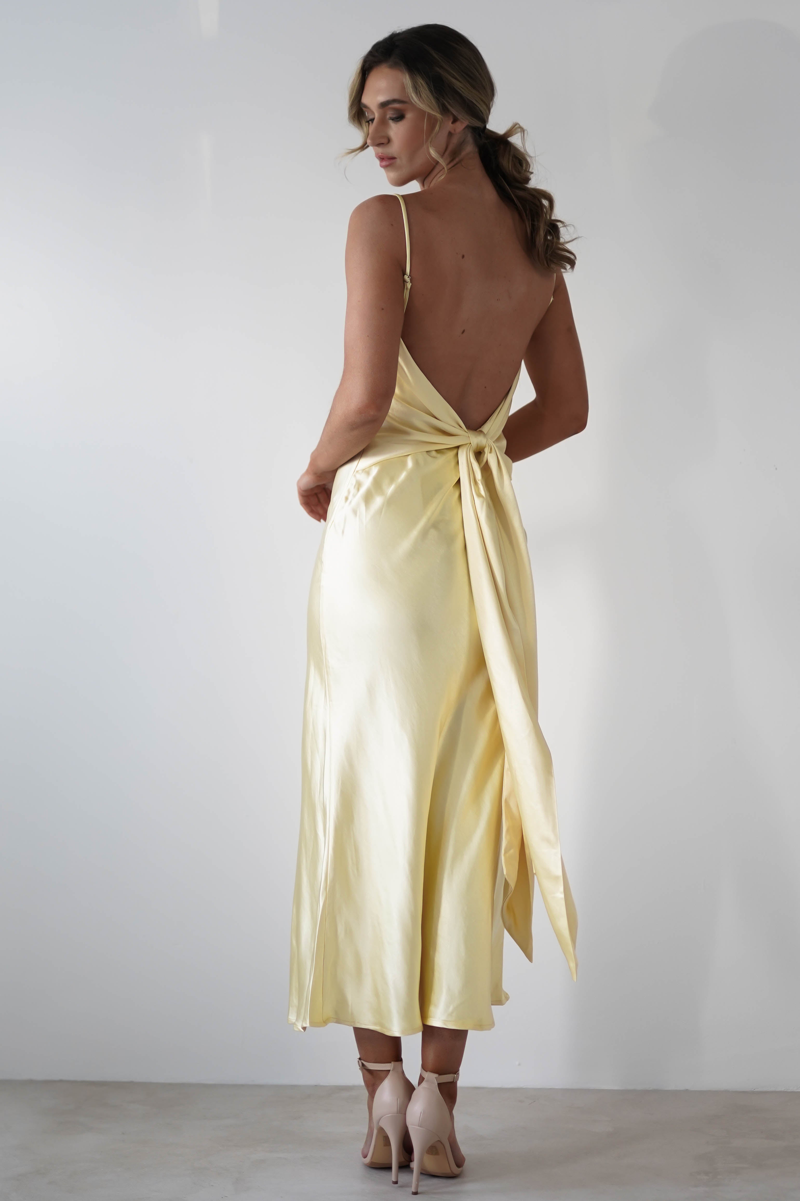 A woman stands facing away, wearing a pale yellow satin knit midi dress with a hidden side zipper and tie at the waist. She has wavy hair, wears high heels, and poses against a plain white background—perfect for bridesmaids.