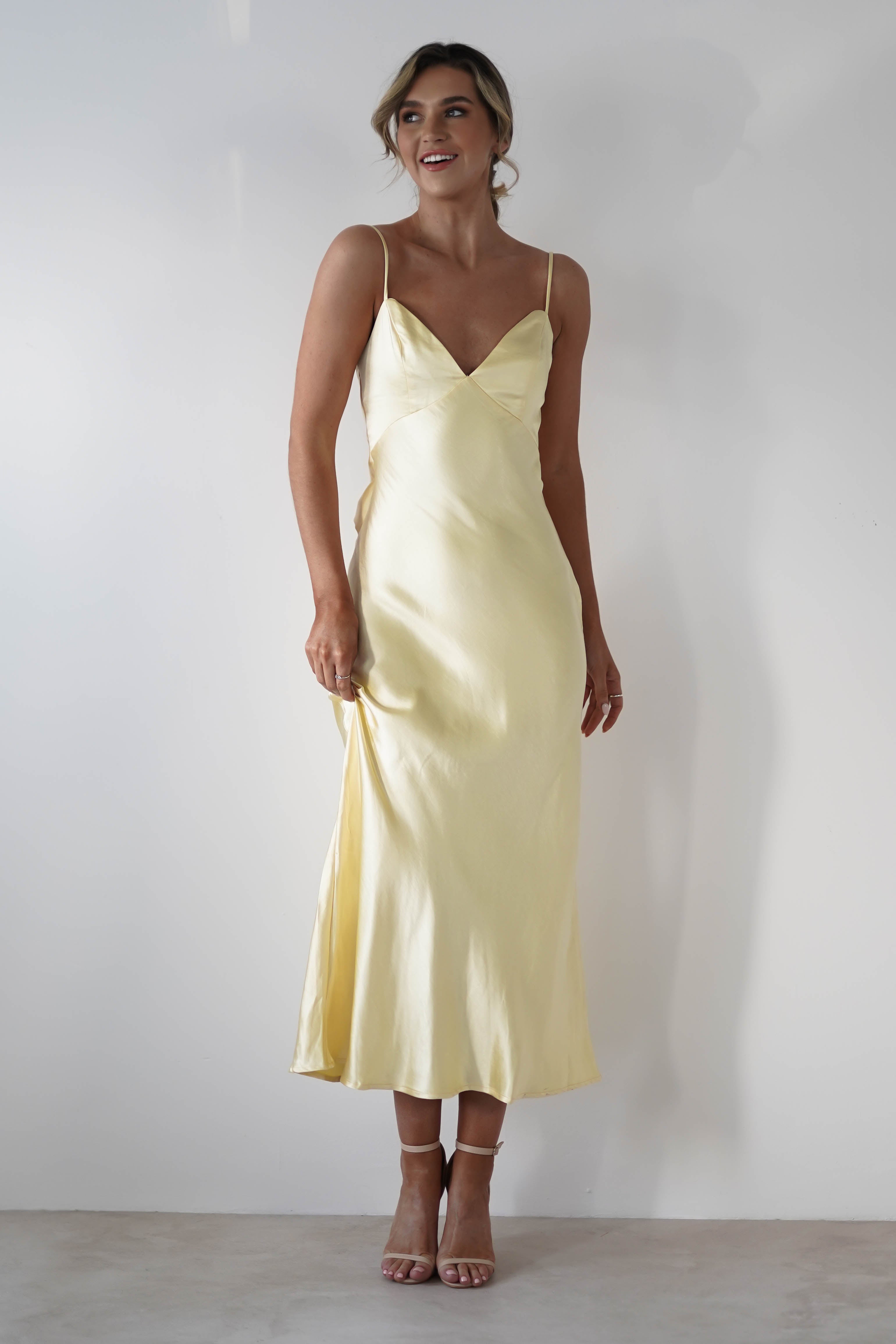A woman in a light yellow satin knit midi slip dress with thin straps and a hidden side zipper stands against a plain white background, looking to the side and smiling. Perfect for bridesmaids, she pairs it with open-toe heels and an elegant updo.