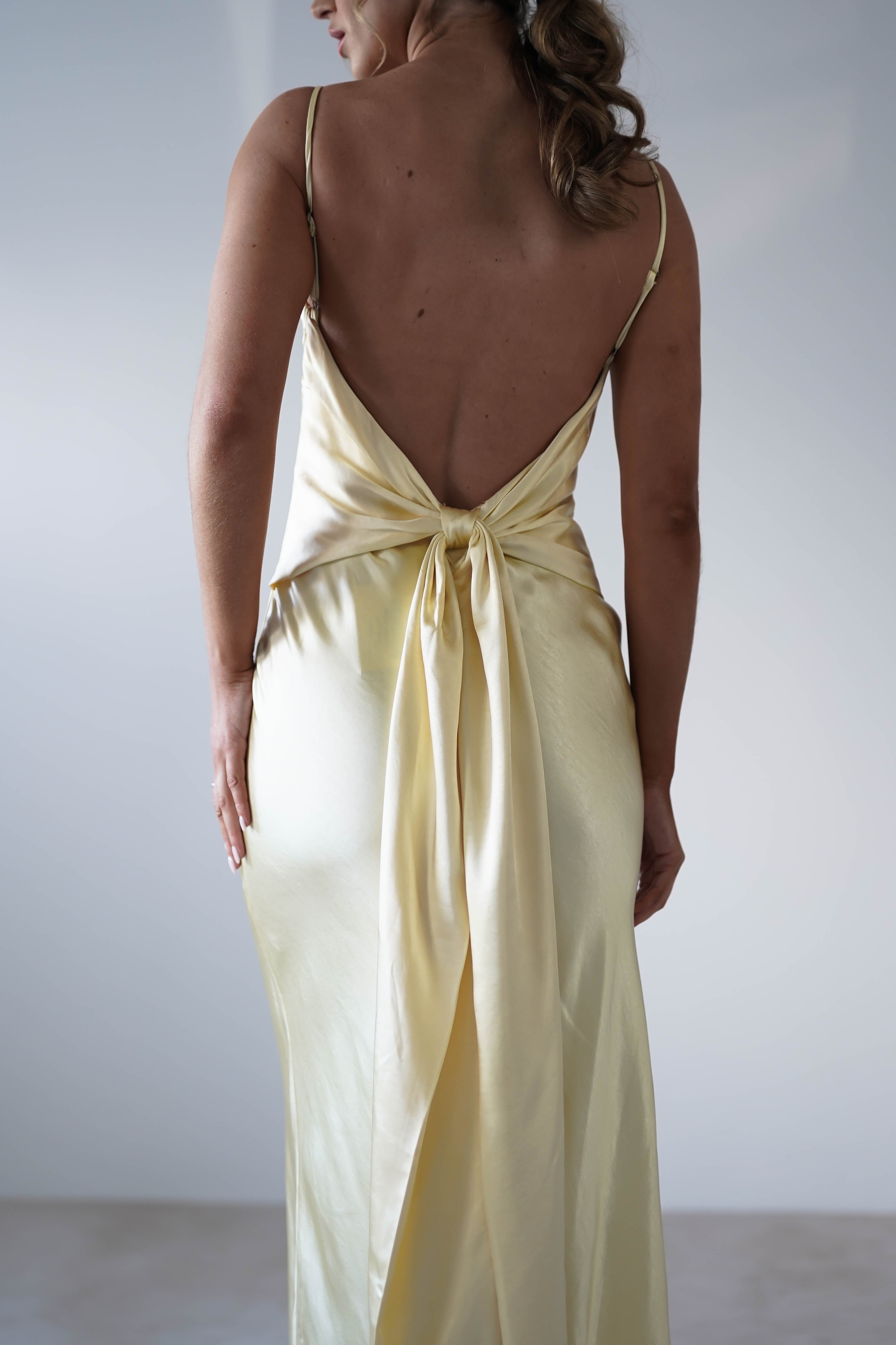 A woman wearing a backless, light yellow satin knit midi dress with spaghetti straps, a sash tied at the waist, and a hidden side zipper—ideal for bridesmaids—photographed from behind against a plain light background.