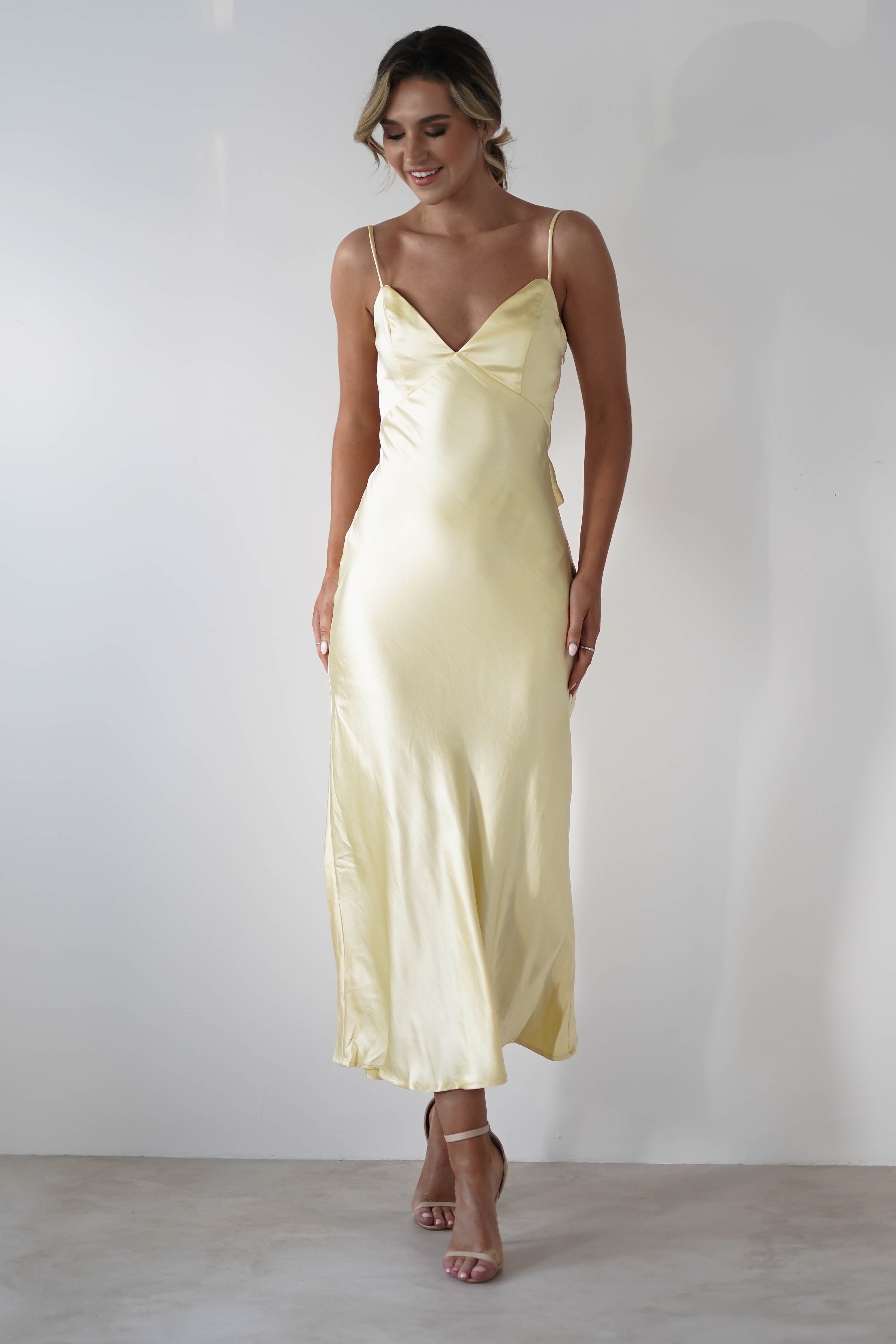 Woman wearing a pale yellow satin knit midi slip dress with thin spaghetti straps, a V-neckline, hidden side zipper, and a flowing ankle-length skirt—perfect for bridesmaids—standing against a plain white background.