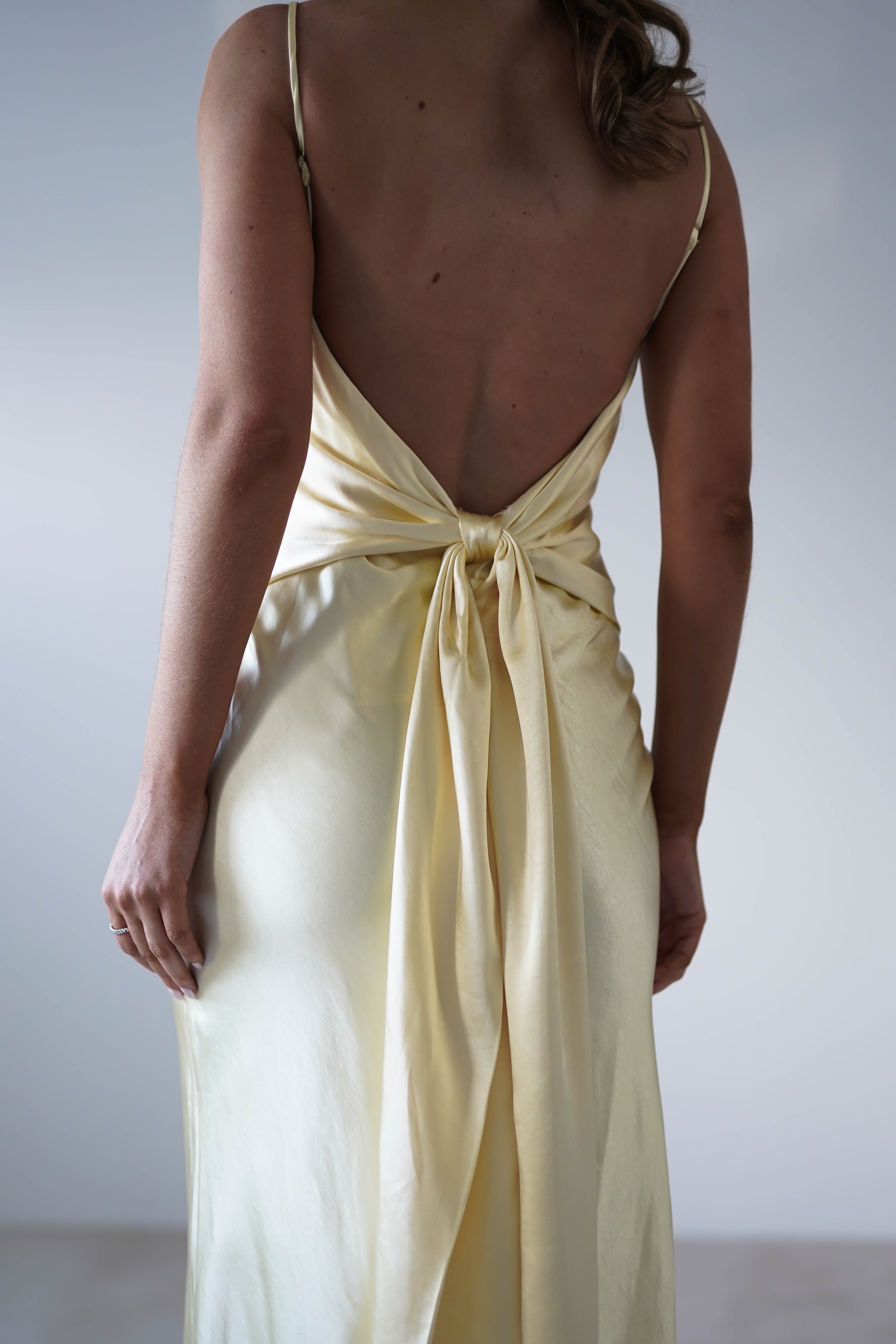 A person wearing a backless, pale yellow satin knit midi dress with a bow at the lower back stands facing away, revealing elegant draping, smooth texture, and a hidden side zipper—perfect for bridesmaids seeking understated sophistication.