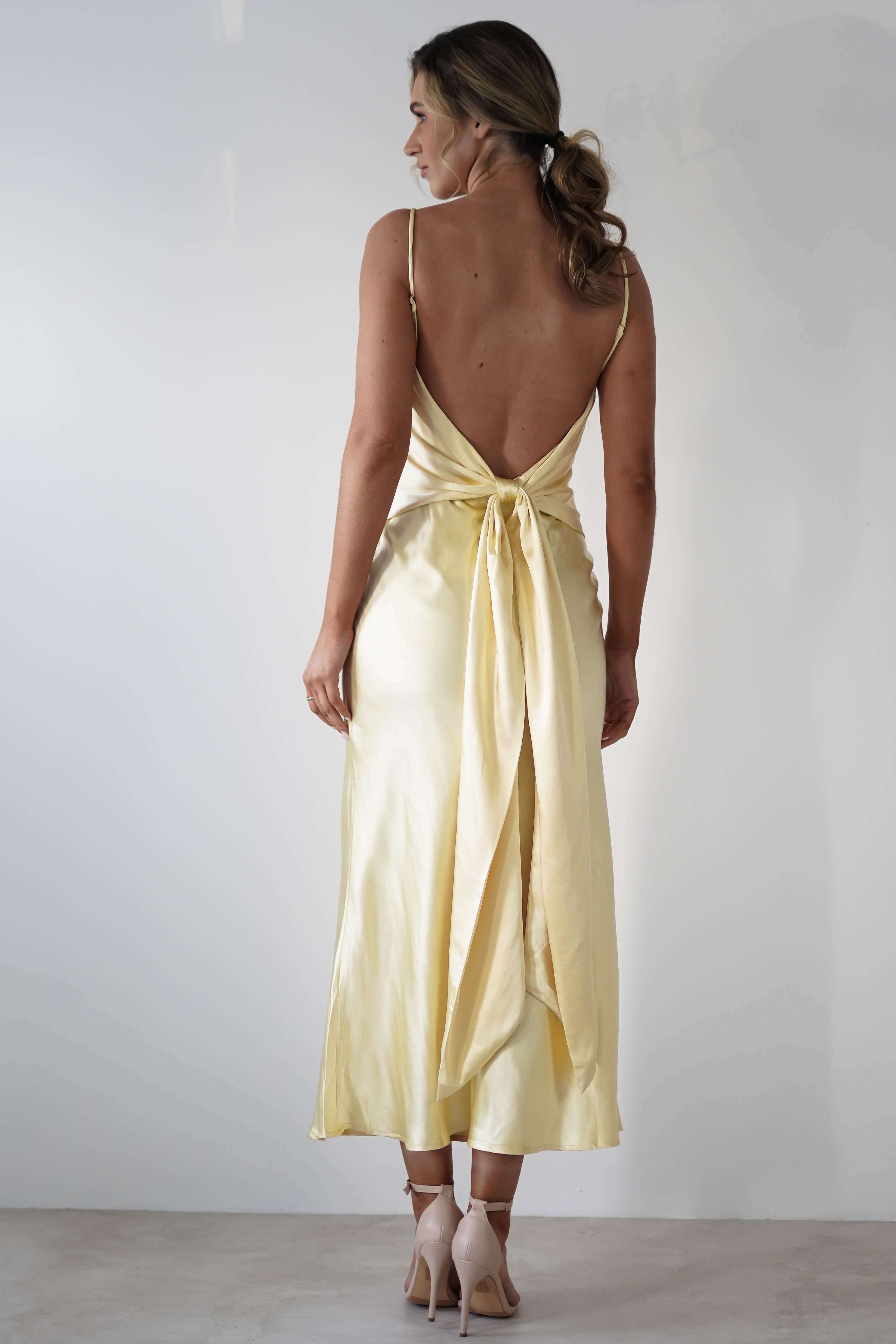 A woman stands facing away, wearing a light yellow satin knit midi dress with thin straps, a large bow at the waist, and a hidden side zipper—an elegant choice for bridesmaids. Her light brown hair is in a loose ponytail and she wears nude heels.