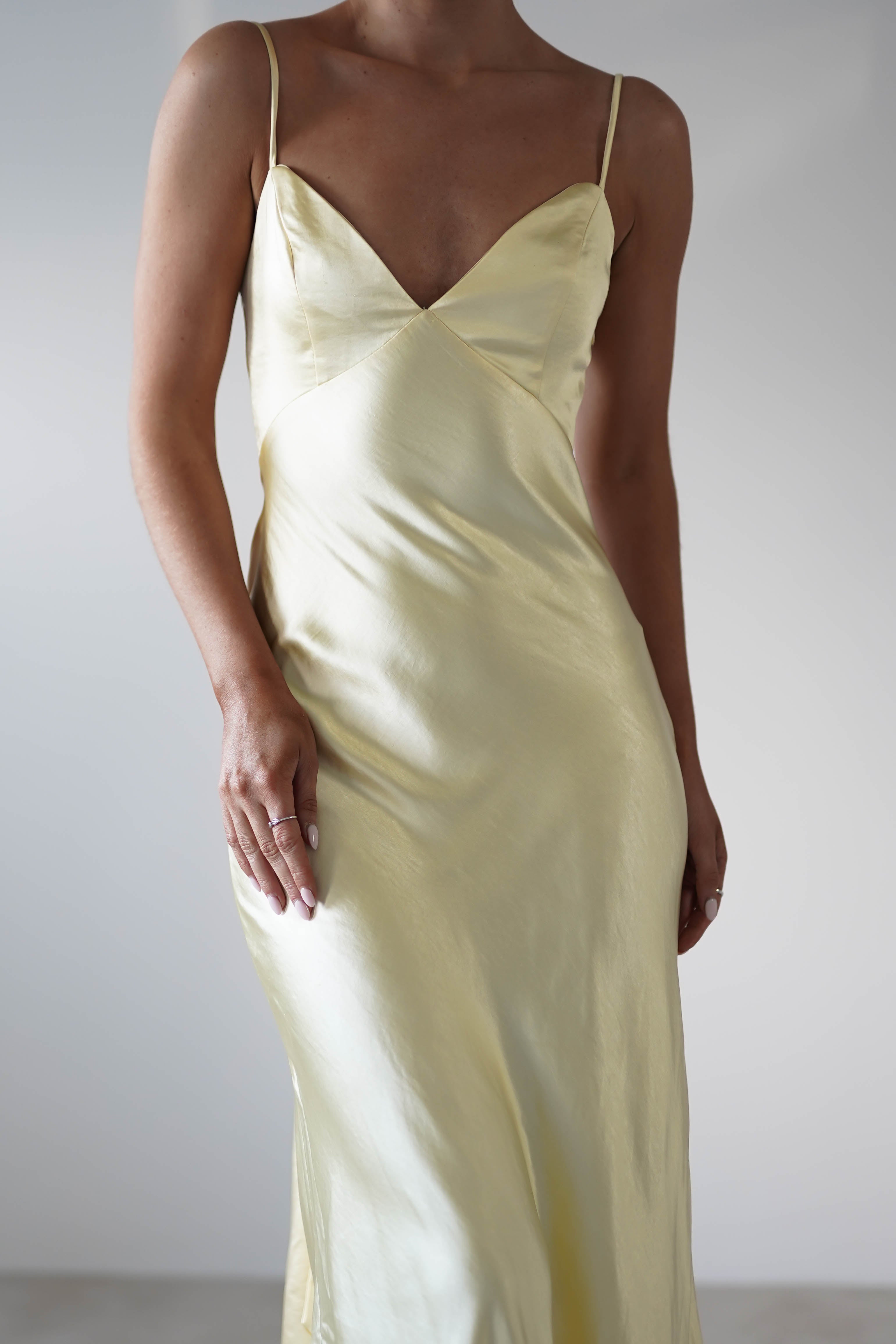 A person wearing a light yellow satin knit midi slip dress with thin straps, a deep V neckline, and a hidden side zipper stands against a plain, light-colored background. Their face is not visible.