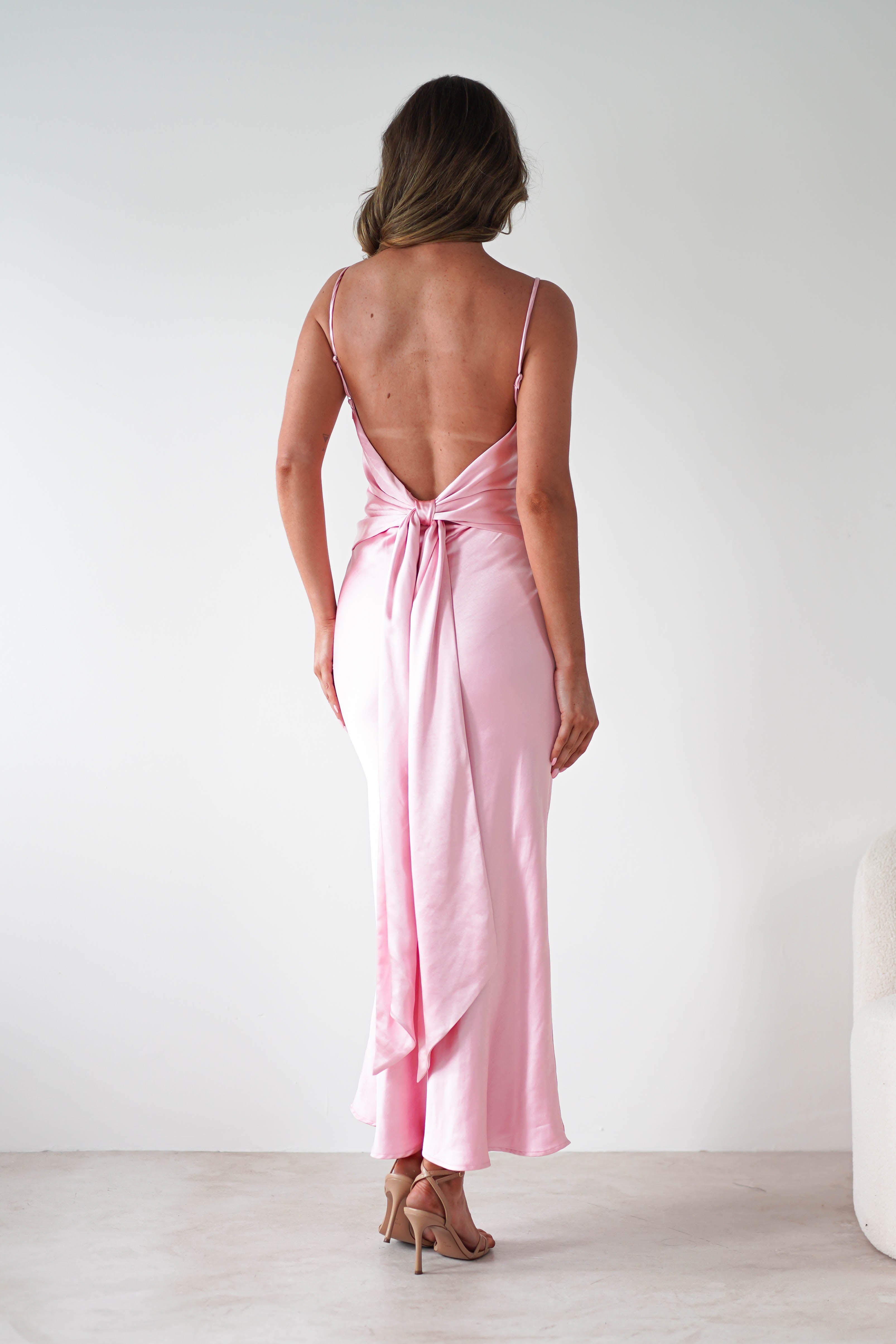 A woman stands facing away, wearing a silky, light pink satin midi dress with thin straps and a bow at the lower back. She is in a minimal, light-colored room and wears nude high heels—a perfect look for bridesmaids.