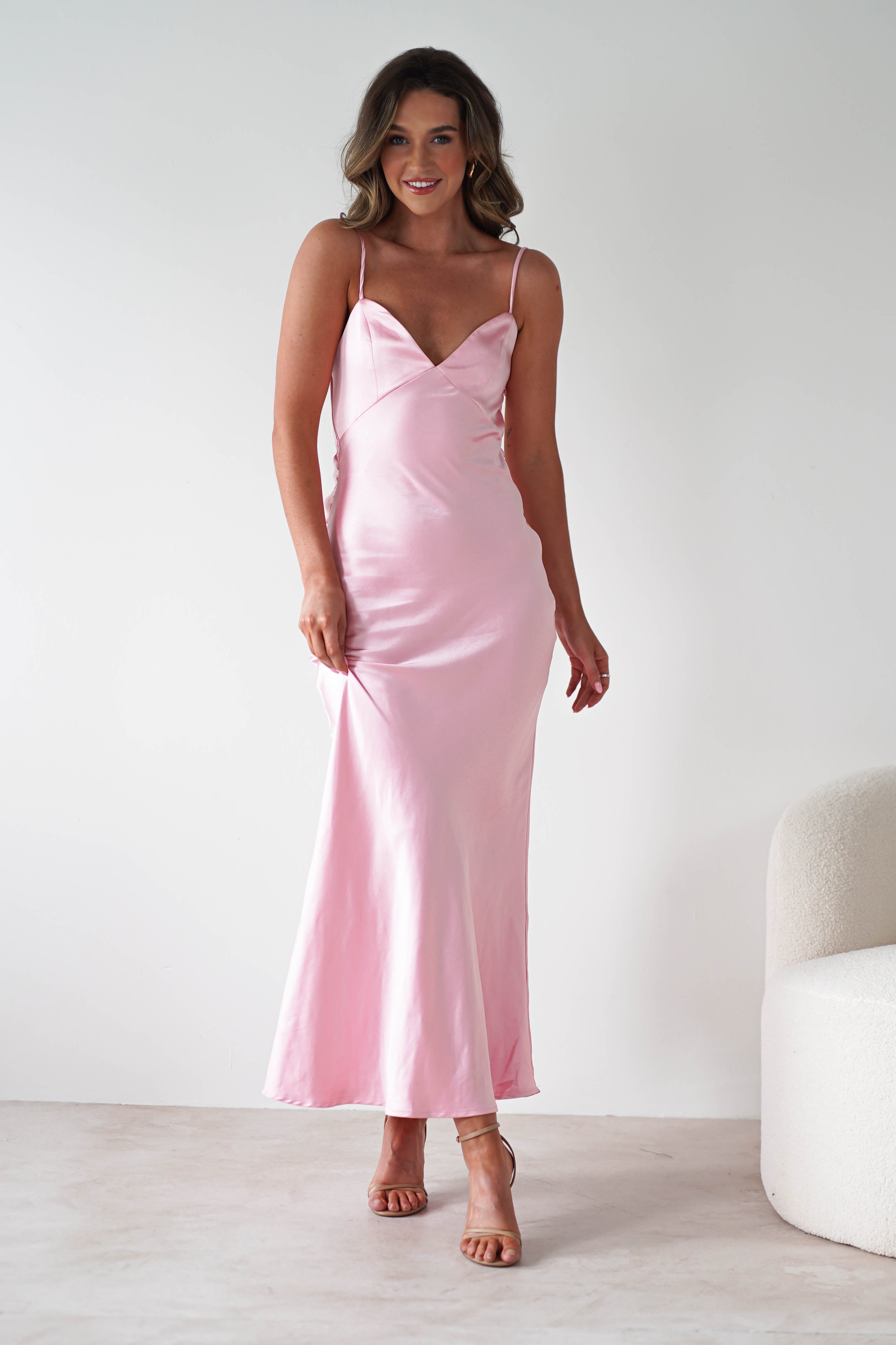 A woman in a light pink satin bridesmaids dress with spaghetti straps stands indoors, smiling and holding the dress. She has wavy, shoulder-length hair and wears nude heels. The background is white and minimalistic.