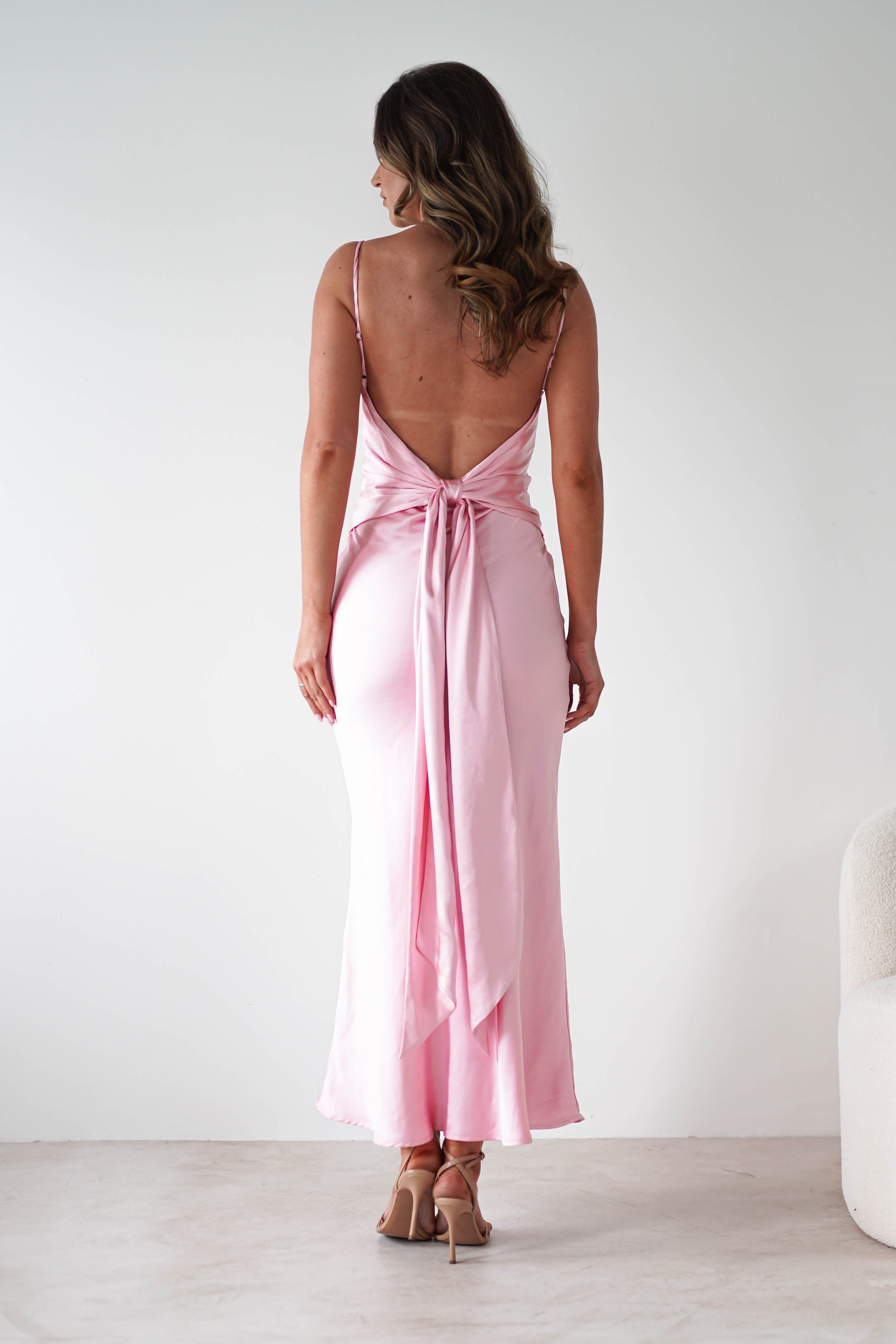 Woman standing with her back to the camera, wearing a pale pink satin midi dress with a bow at the waist. She has long, wavy brown hair and beige high heels, set against a plain white background. Perfect as an elegant bridesmaids dress.