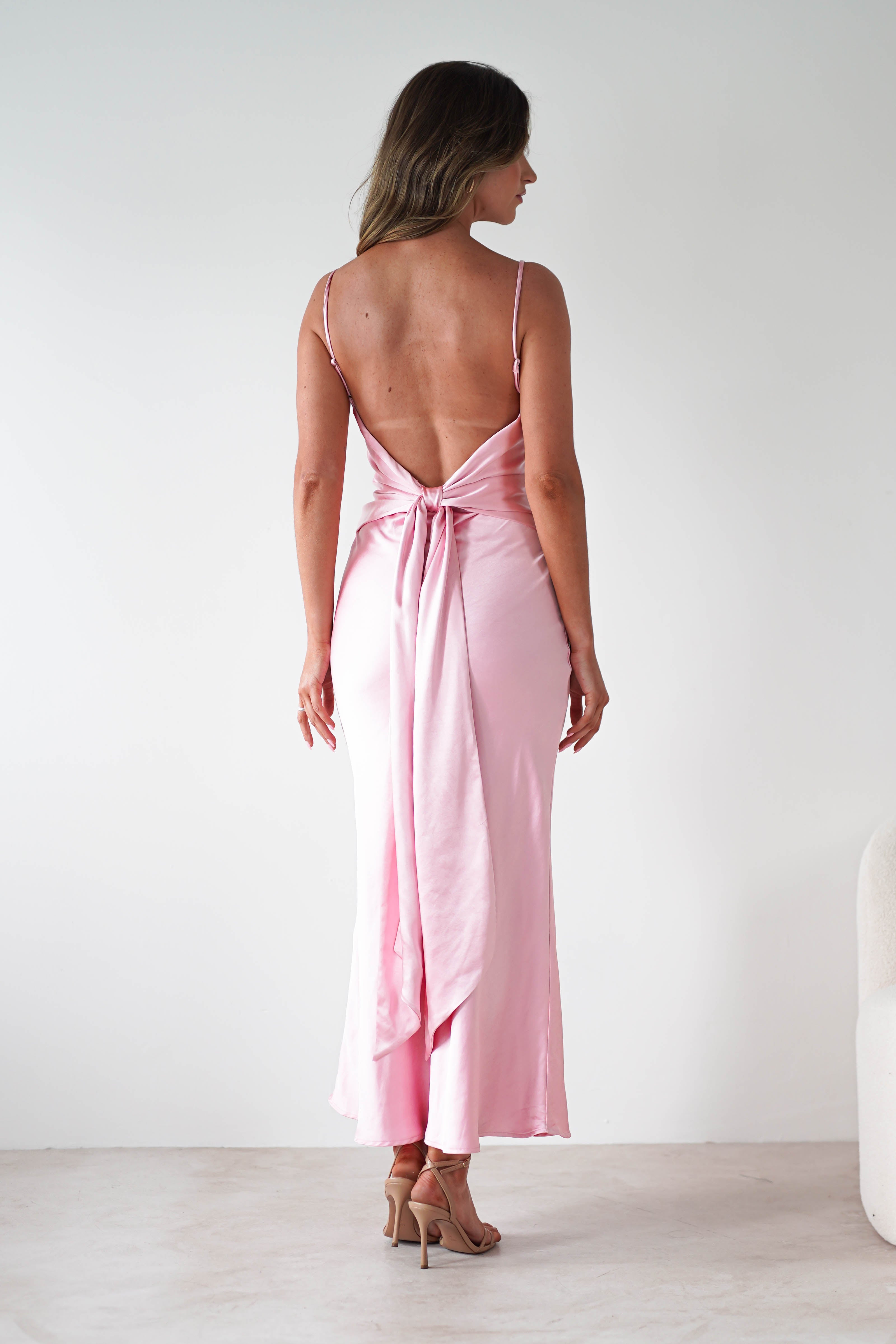 A woman stands facing away, wearing a light pink satin bridesmaids dress with thin straps, an open back, and a large bow at the waist. The elegant midi dress flows to her calves as she poses on a light floor against a plain white background.