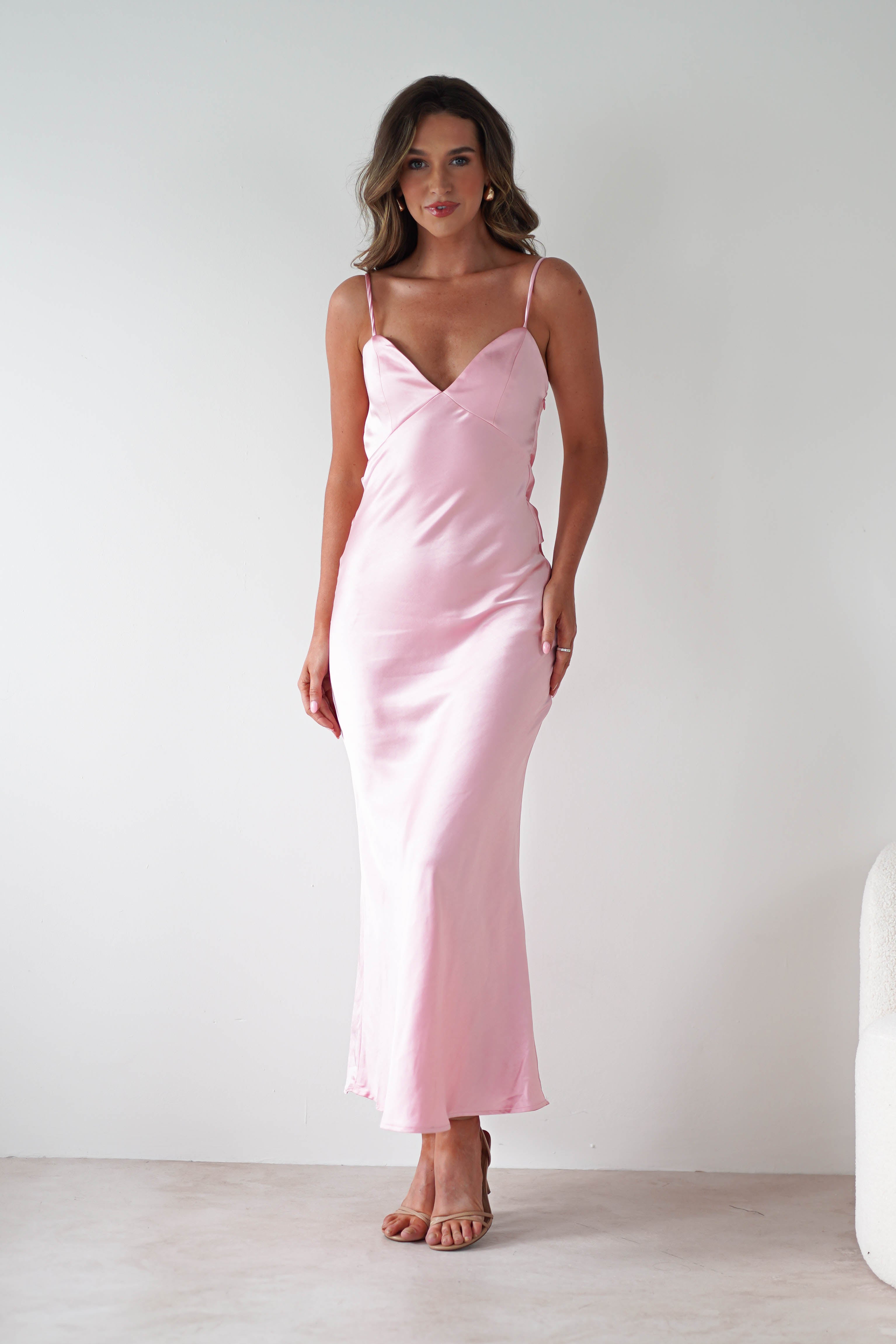 A woman stands against a plain white background wearing a long, light pink satin bridesmaids dress with thin straps and a V-neckline. She has loose, wavy hair and is posing with a slight smile.
