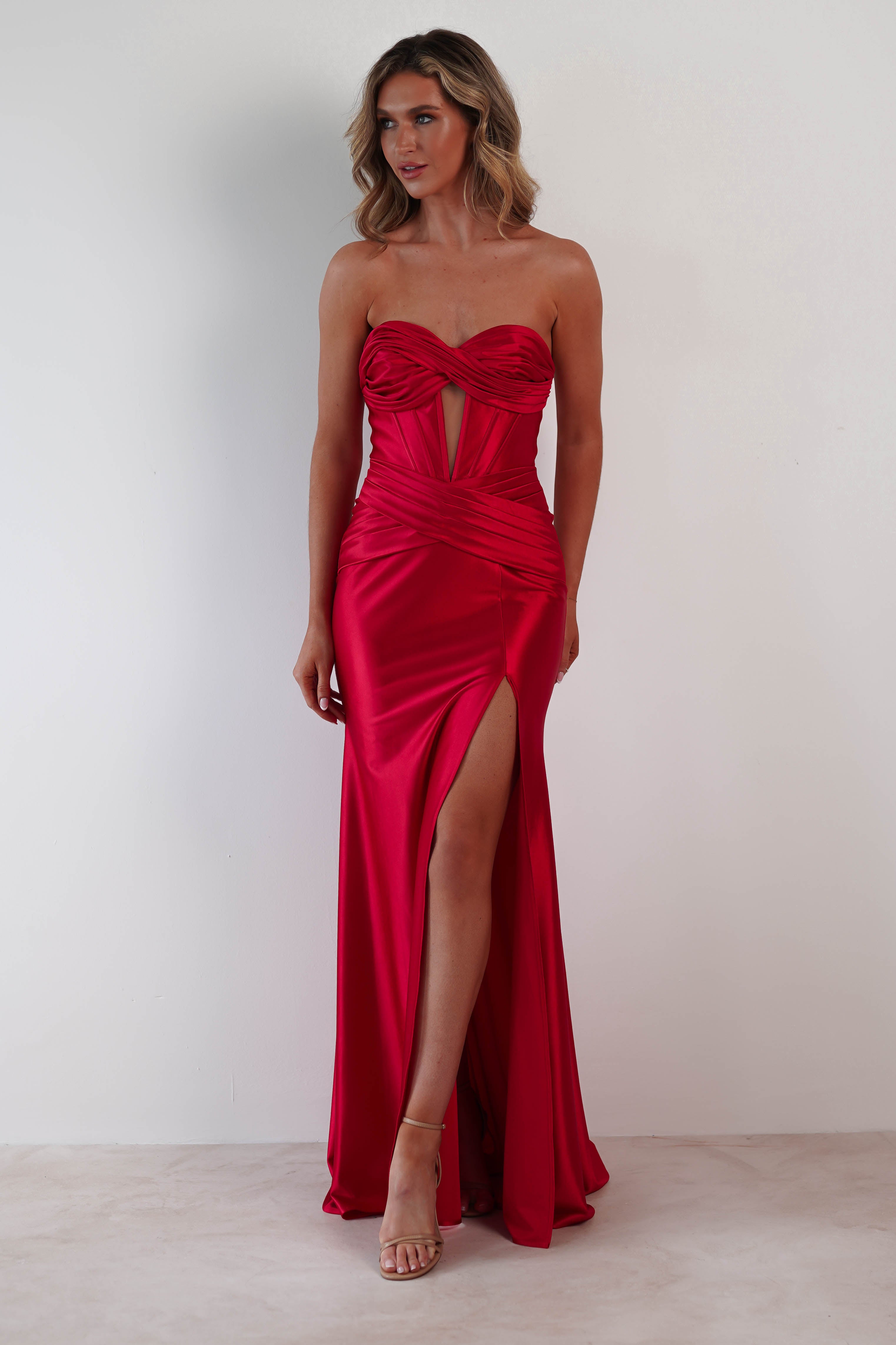A woman in a strapless satin prom dress stands against a white background. The red gown boasts a sweetheart neckline, ruched details, and a high slit on the left side. Her long, wavy hair complements her elegant high heels perfectly.