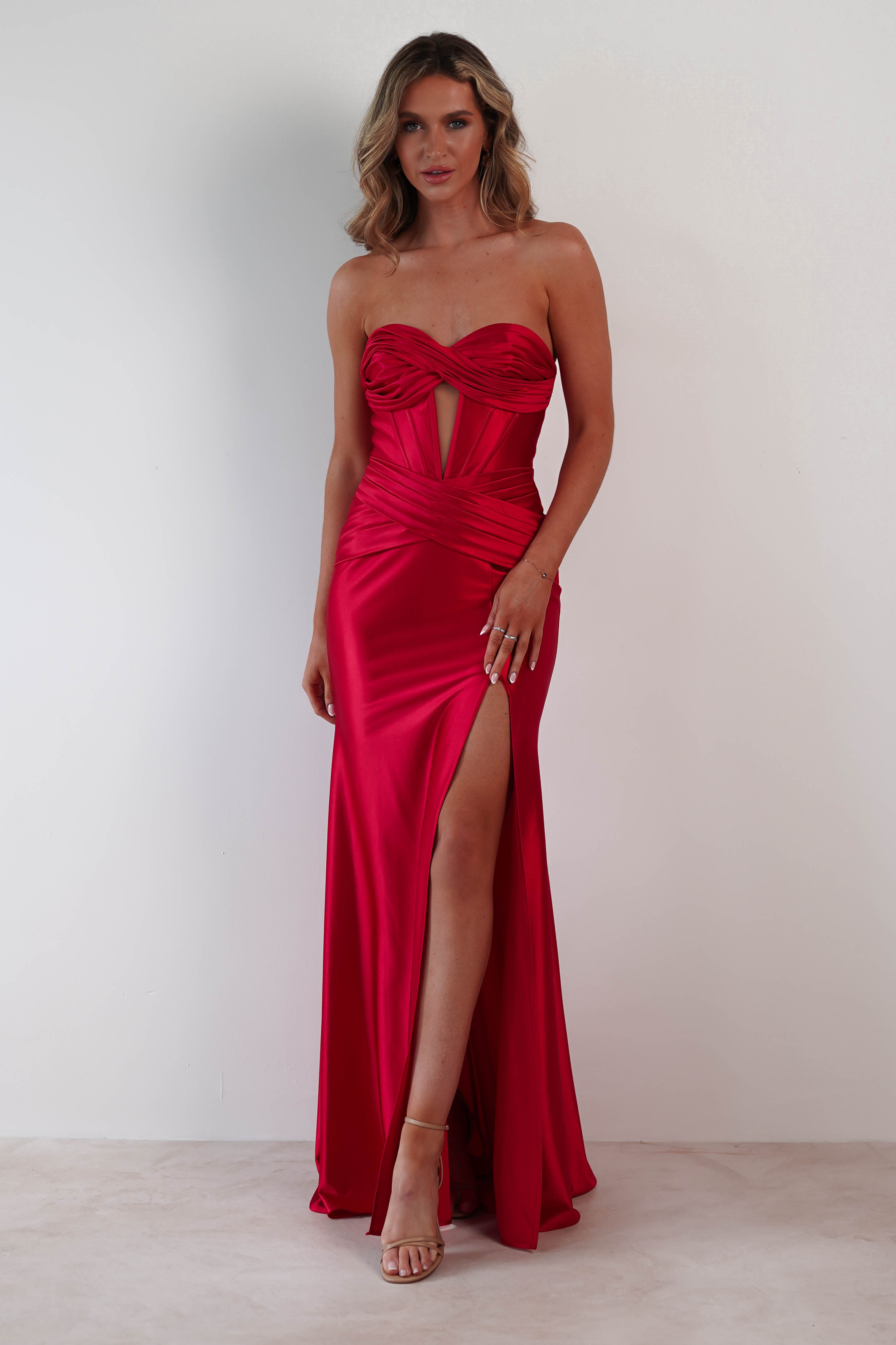 A person wearing a strapless red satin gown with a thigh-high slit stands against a plain white background. The prom dress features ruched detailing and cutouts on the bodice. They have medium-length hair and are wearing strappy heels.
