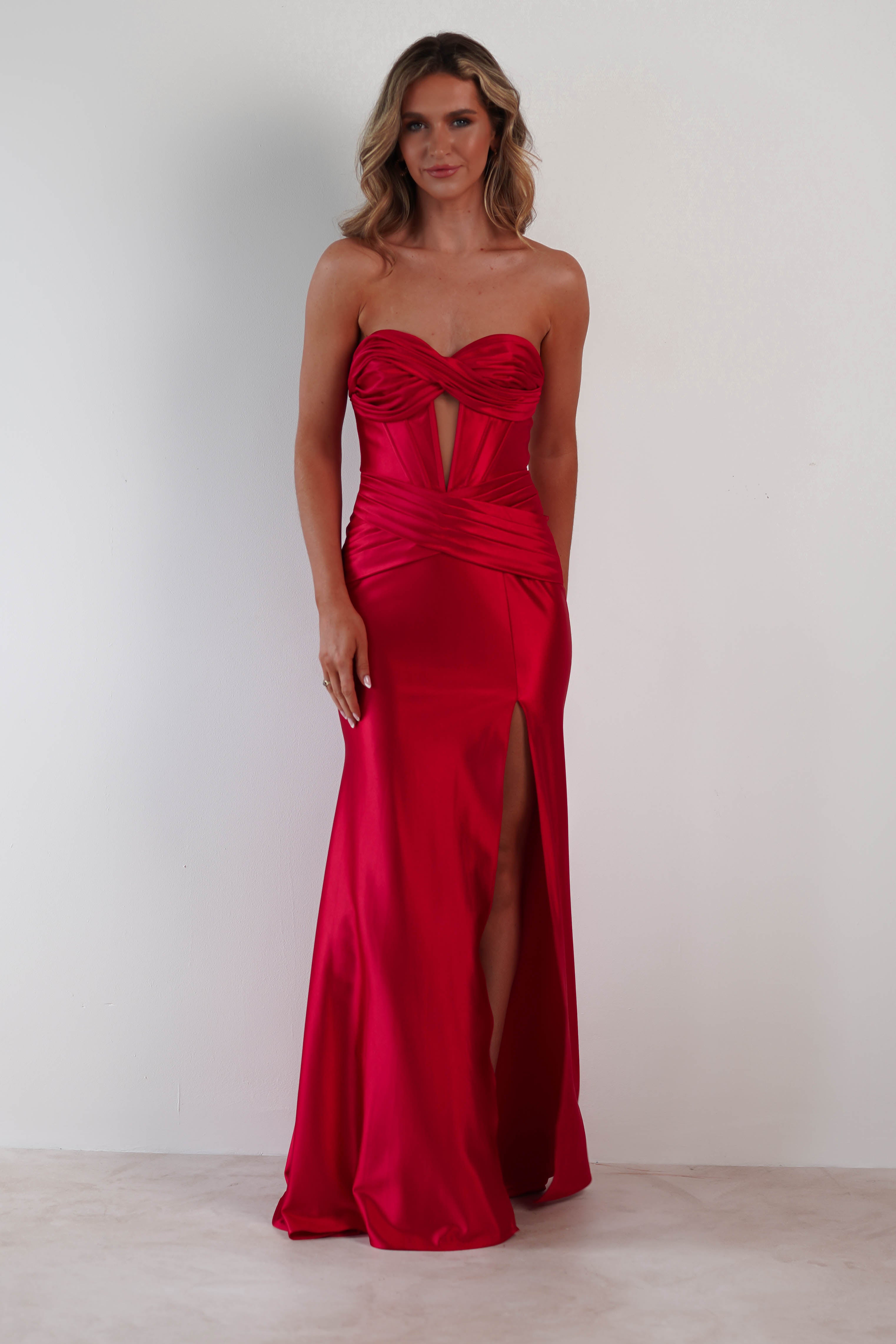 A woman in a strapless, fitted red gown with ruched detailing stands against a plain white background. Crafted from lustrous satin fabric, the dress features a small front cutout and a thigh-high slit. She has long, wavy hair and a neutral expression.