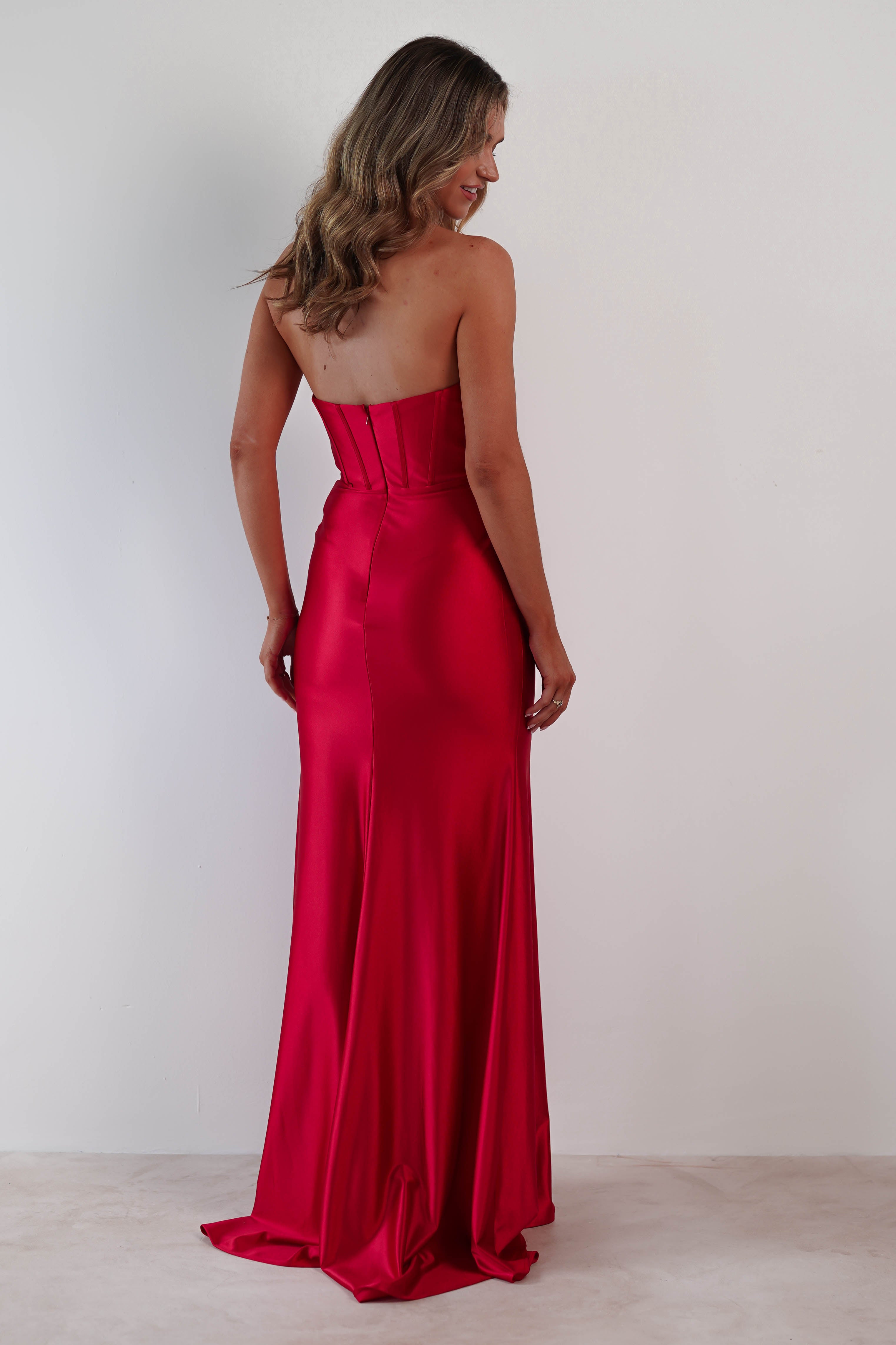 A woman stands with her back to the camera, wearing a strapless prom dress in stunning red satin fabric. Her wavy hair cascades past her shoulders, complementing the gowns fitted bodice and flared skirt against a plain white background.