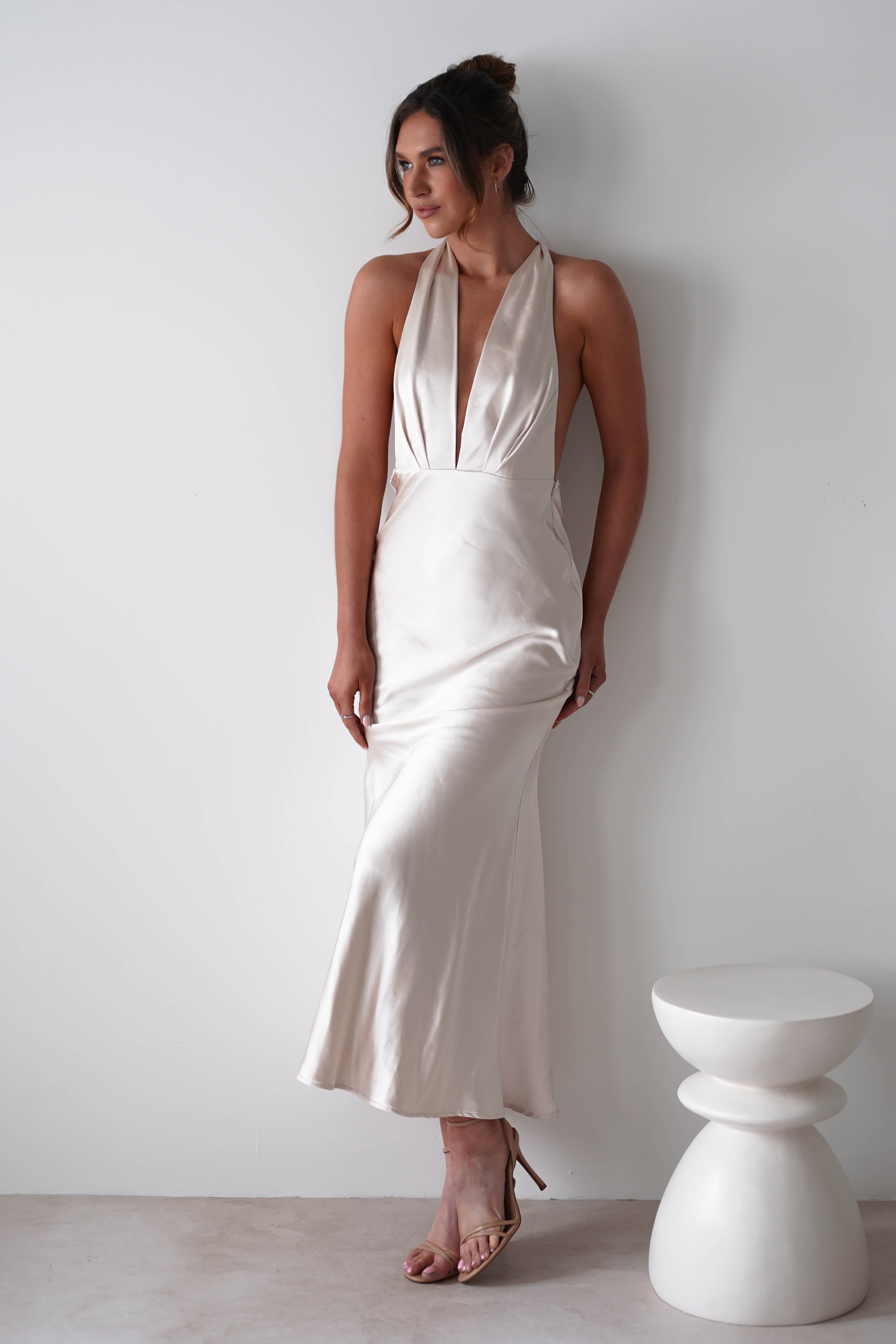 A woman in a sleeveless, ivory soft satin material halter dress with a deep V-neckline stands by a white wall next to a modern sculptural stool—perfect for rehearsal dinners, with strappy nude heels and her hair styled in a loose updo.